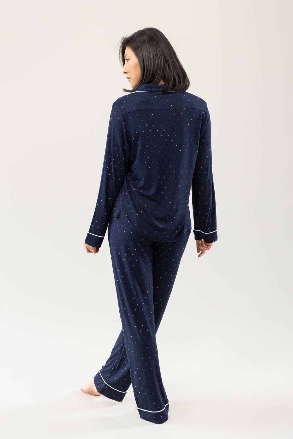 A person with shoulder-length hair stands barefoot, facing away, wearing the Cozy Earth Women's Bamboo Stretch-Knit Long Sleeve Pajama Set in navy blue with small white polka dots and light-colored piping on the cuffs against a plain white background. 