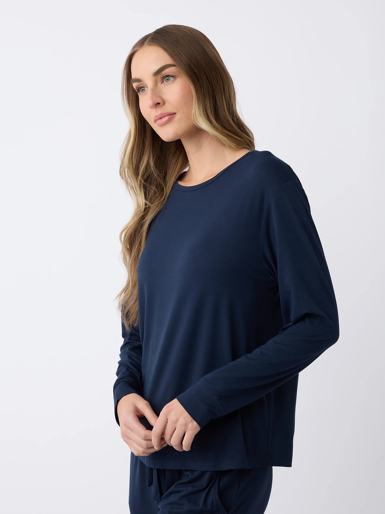 With long hair, someone wears a Cozy Earth Women's Stretch Knit Bamboo Long Sleeve Lounge Tee in navy, paired with matching pants. They stand against a plain, light background, gazing calmly to the side. |Color:Navy