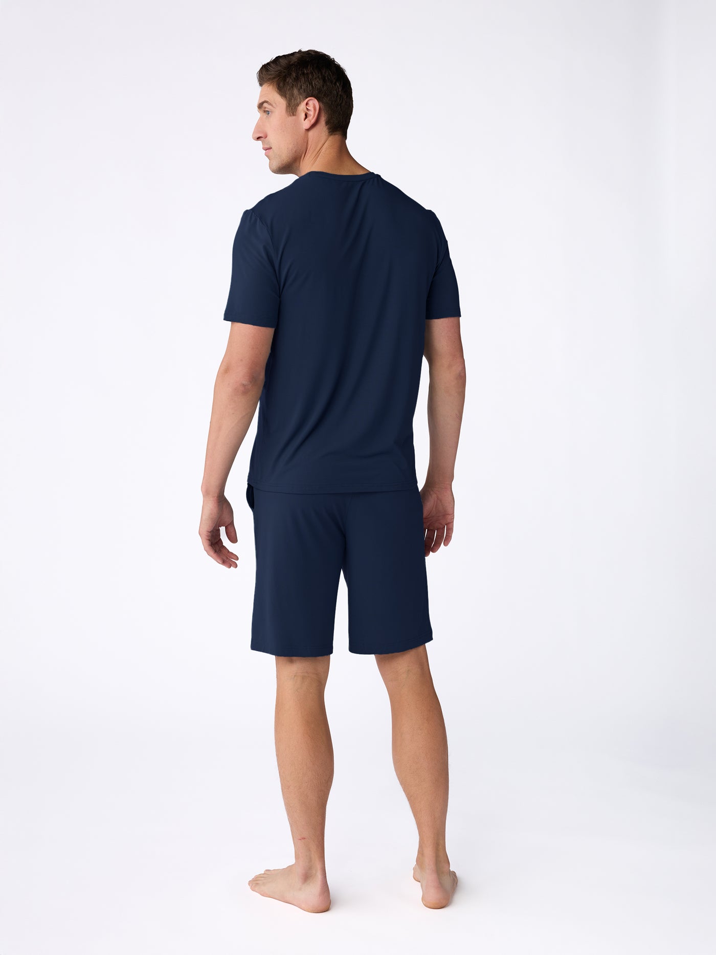 A man stands barefoot on a white background, facing away from the camera, wearing the Cozy Earth Men's Bamboo Stretch-Knit Short Sleeve Pajama Set in navy blue. 