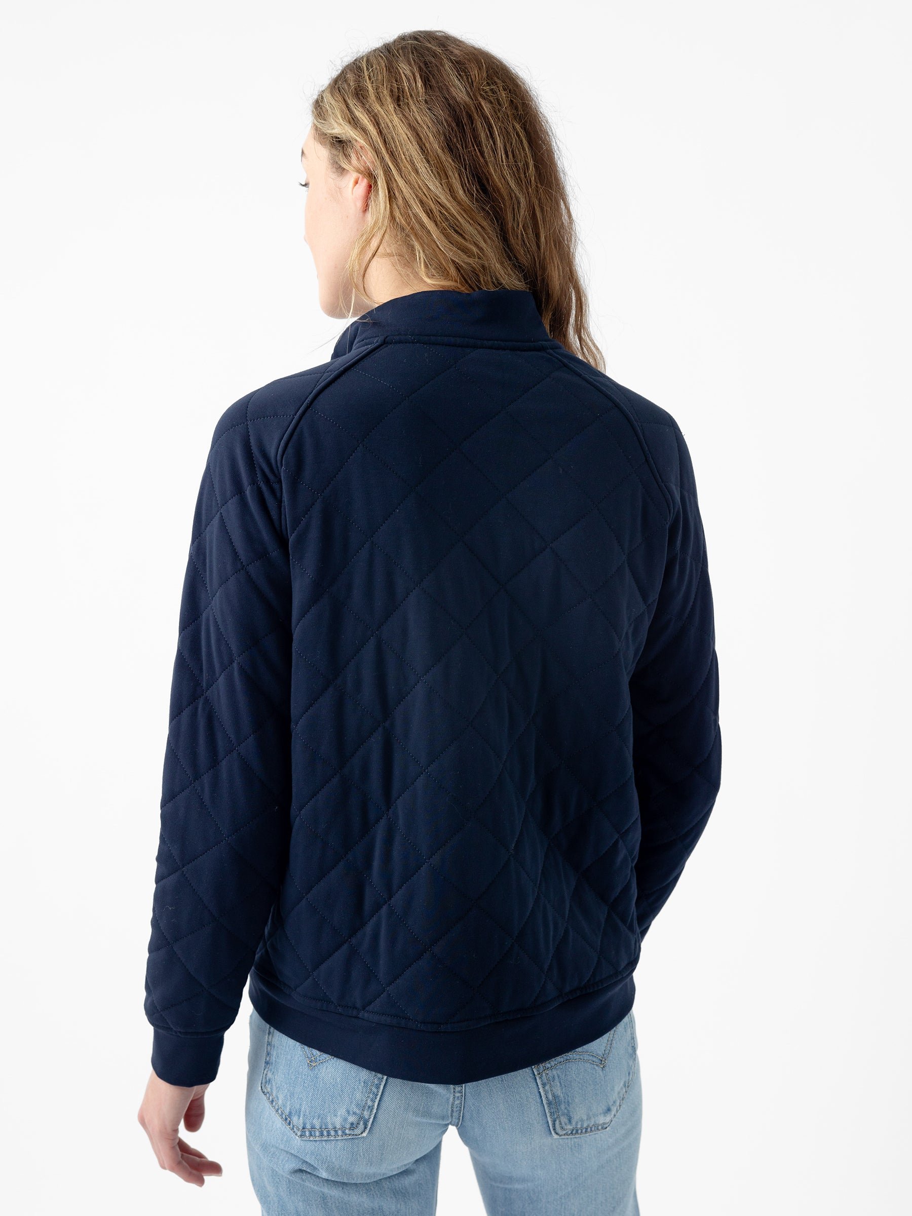 A person with long, wavy hair wearing a dark blue Women's Ultra-Soft Bamboo Quilted Snap Pullover from Cozy Earth and light blue jeans stands facing away from the camera. The background is plain white. 