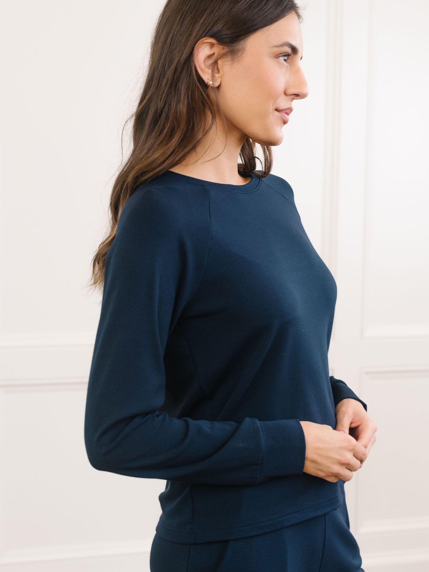 A woman with long dark hair stands in profile against a white background, wearing Cozy Earth's navy blue fitted, long-sleeve pullover from their Ultra-Soft Bamboo Pant & Crew Set. 