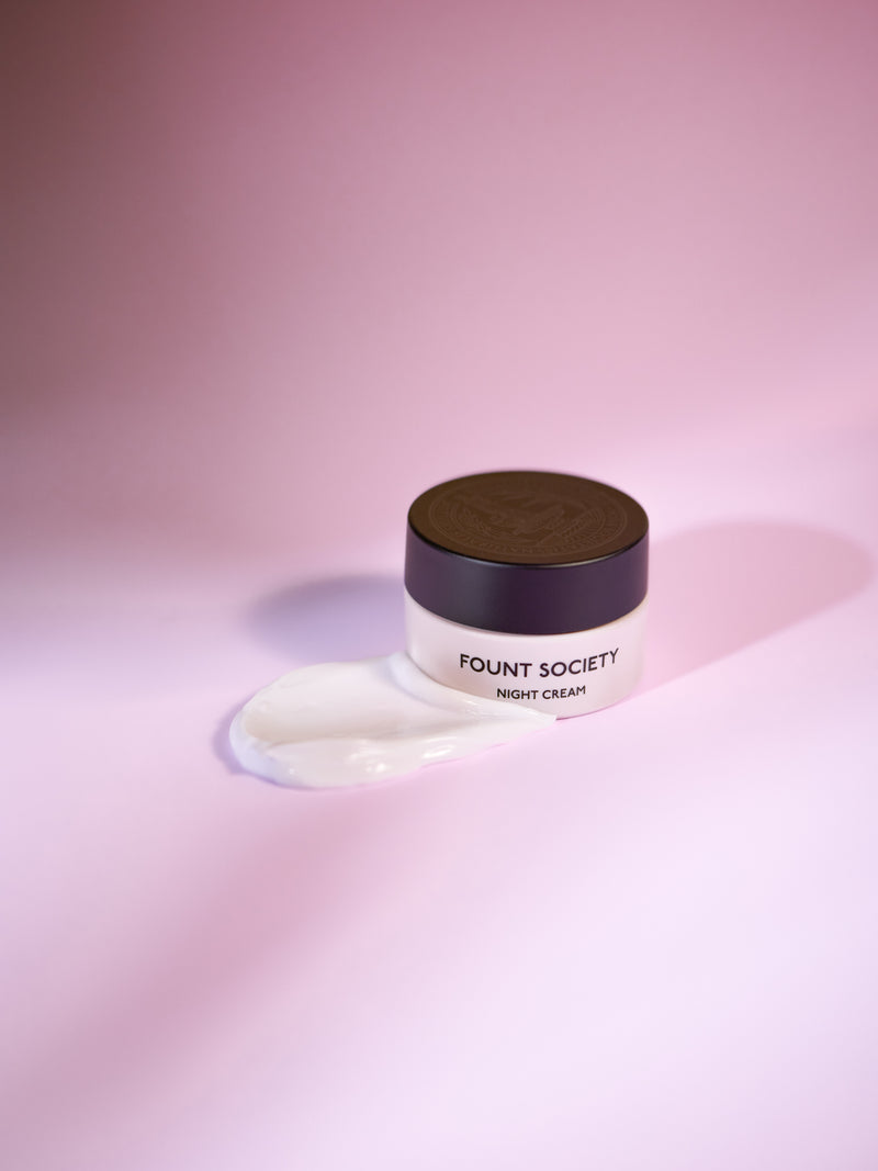 A jar of Cozy Earth Night Cream sits on a pink surface, with some of the cream spilled beside it. The jar features a dark lid that stands out against the soft background.