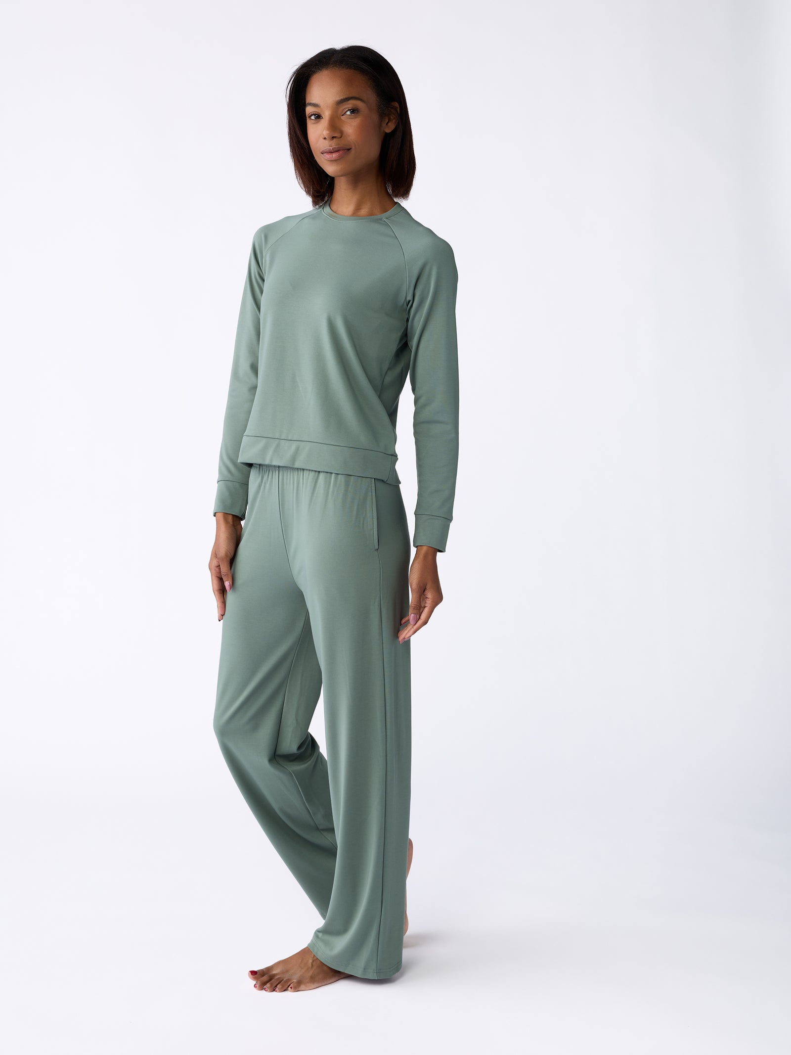 A person stands barefoot against a plain white background, wearing the Women's Ultra-Soft Bamboo Pullover Crew by Cozy Earth in a matching sage green loungewear set, featuring a long-sleeve top and wide-leg pants. 