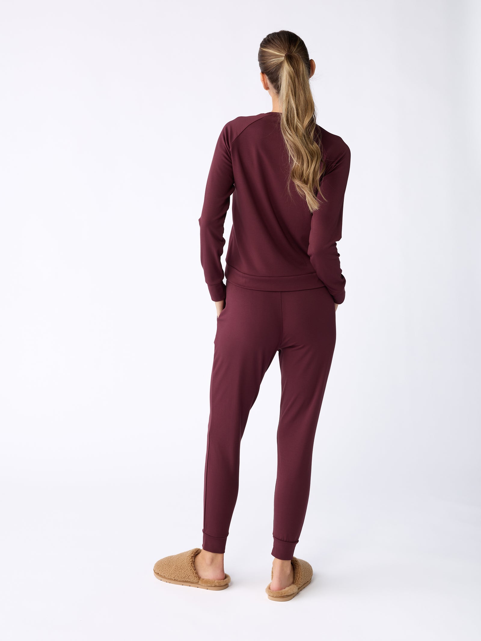 A person stands facing away, dressed in a matching set featuring the Women's Ultra-Soft Bamboo Pullover Crew from Cozy Earth in burgundy, paired with tan slippers. Their long hair is tied in a ponytail against a plain, light-colored background. 