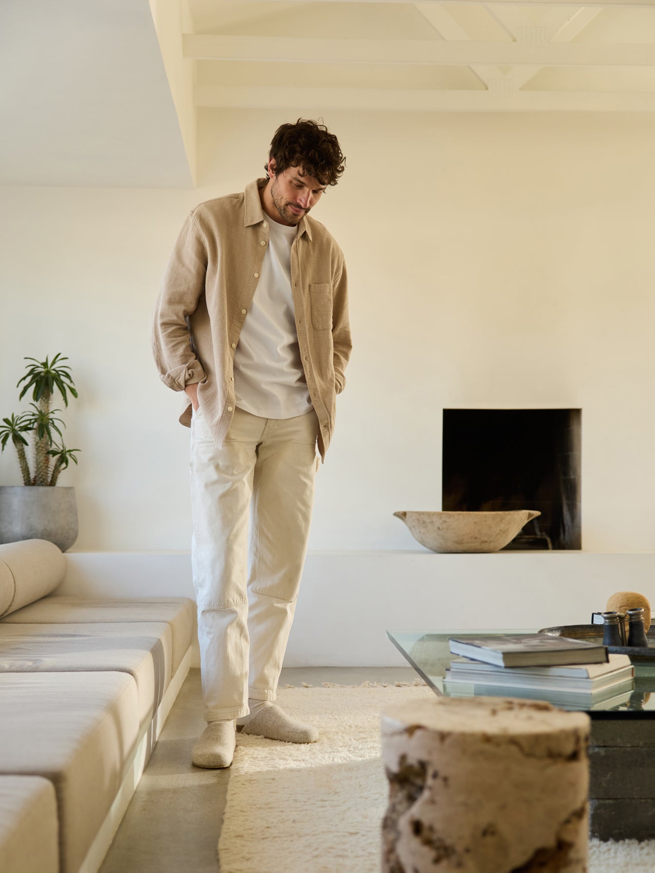 A curly-haired man stands in a minimalist living room, wearing a beige shirt, white t-shirt, light pants, and Cozy Earth's Lakehouse Clog. The room features a sofa, glass coffee table, plants, and a fireplace with a large bowl on the hearth. 