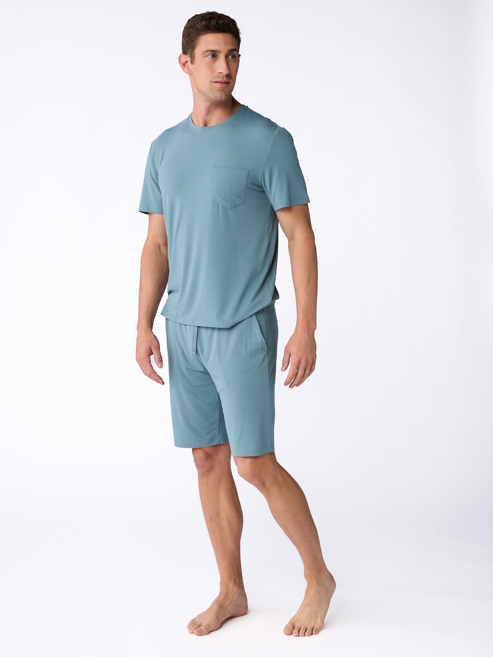 A man stands barefoot wearing Cozy Earth's light blue Men's Stretch-Knit Bamboo Pajama Short. The T-shirt has a pocket on the left side. He is gazing to the right, with his arms relaxed by his sides, set against a plain white background. 