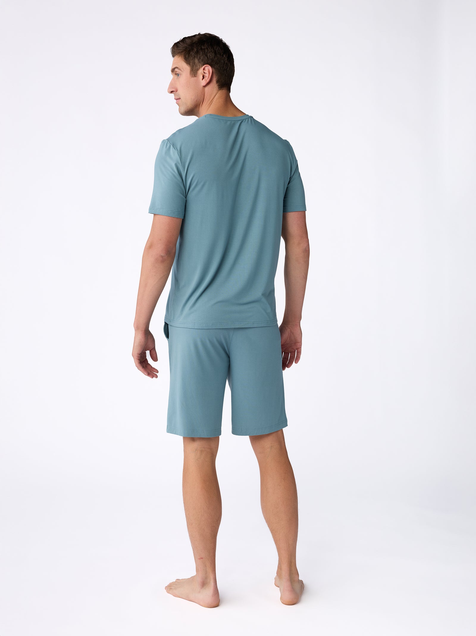 A man stands barefoot, facing left, in a minimalistic setting, wearing the Men's Stretch-Knit Bamboo Lounge Tee from Cozy Earth in a light teal color. The plain white background highlights this casual outfit. 