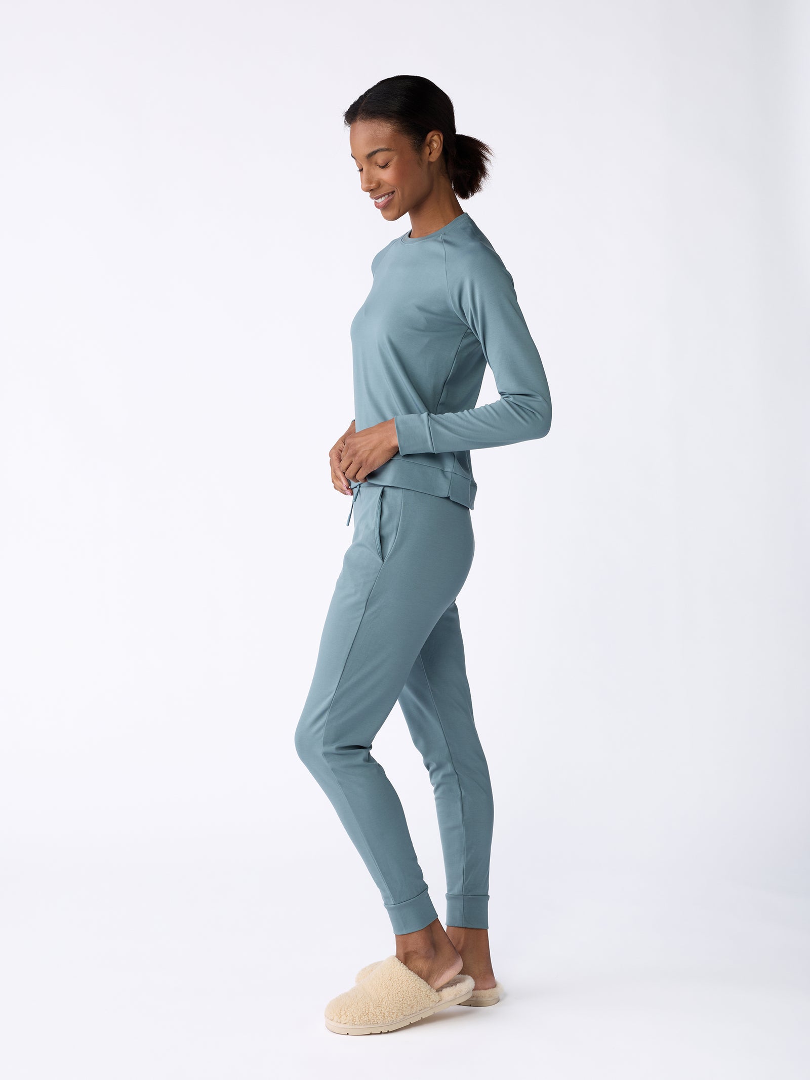 A woman wearing Cozy Earth's Women's Bamboo Jogger Pant in teal and beige slippers smiles while standing in a relaxed pose against a white background. Her hair is pulled back as she stands in profile, with her hands resting on her hips. 