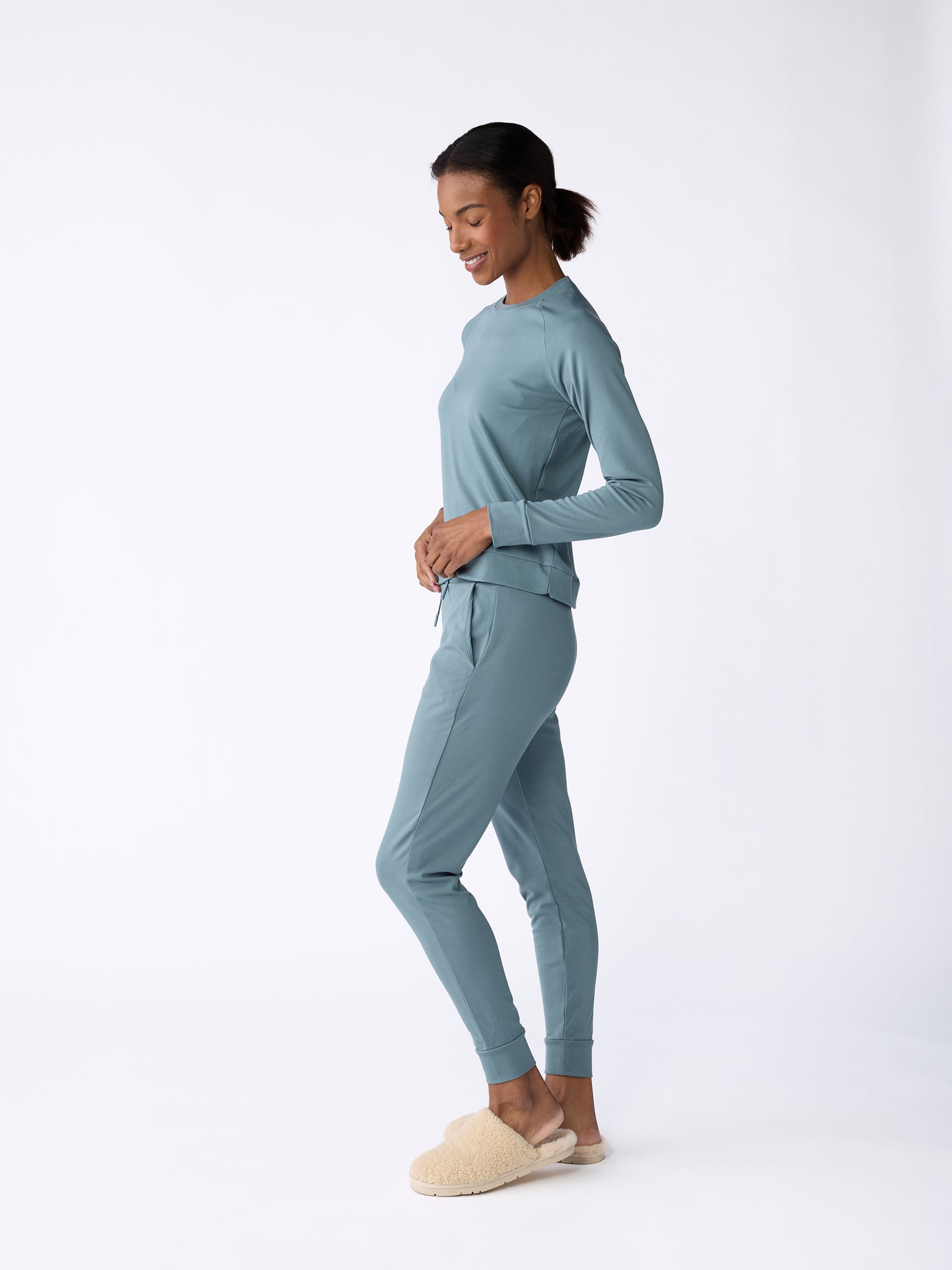 A person in a relaxed pose wearing the Women's Ultra-Soft Bamboo Pullover Crew by Cozy Earth in light blue, paired with matching jogger pants and cream-colored slippers. The background is plain white. 