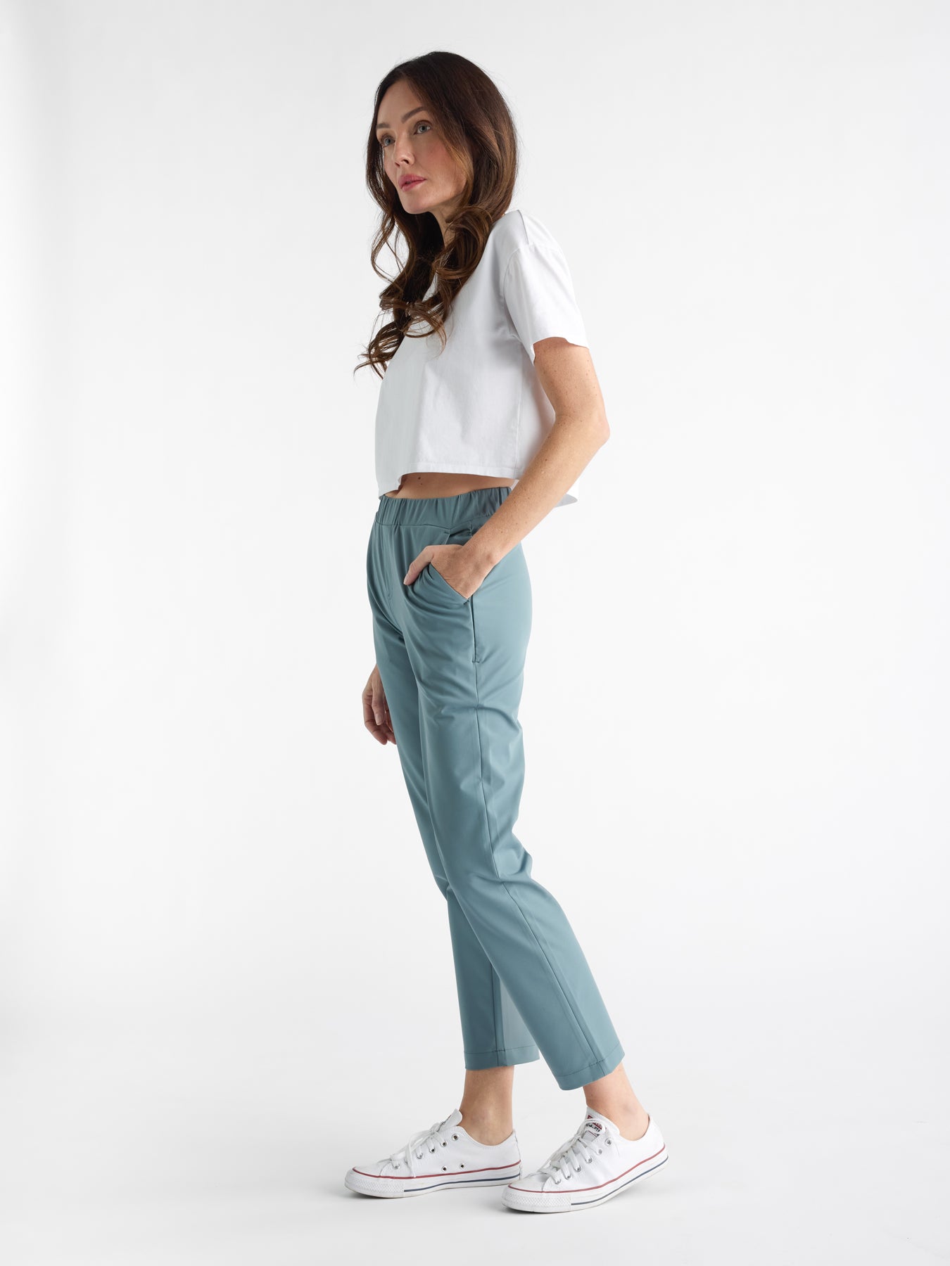 Standing in profile against a white background, a person with long hair wears a white cropped T-shirt, teal Women's Always Cropped Pant by Cozy Earth, and white sneakers. With one hand in a pocket, they gaze into the distance. 