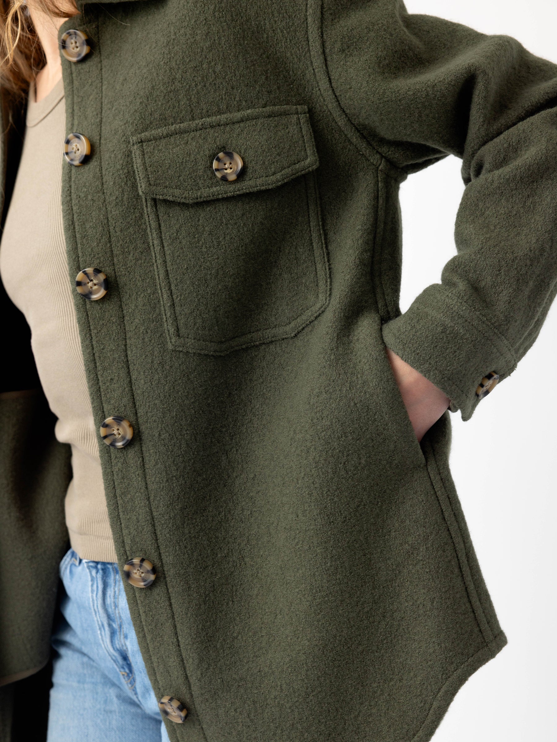 A person wearing the Cozy Earth Women's Boucle Shacket, a dark green button-up jacket featuring light brown buttons and a large front pocket. Complementing this, they are dressed in a beige shirt and blue jeans, with one hand casually placed in the shacket's side pocket. Only a portion of the person is visible in the image. |Color:Olive