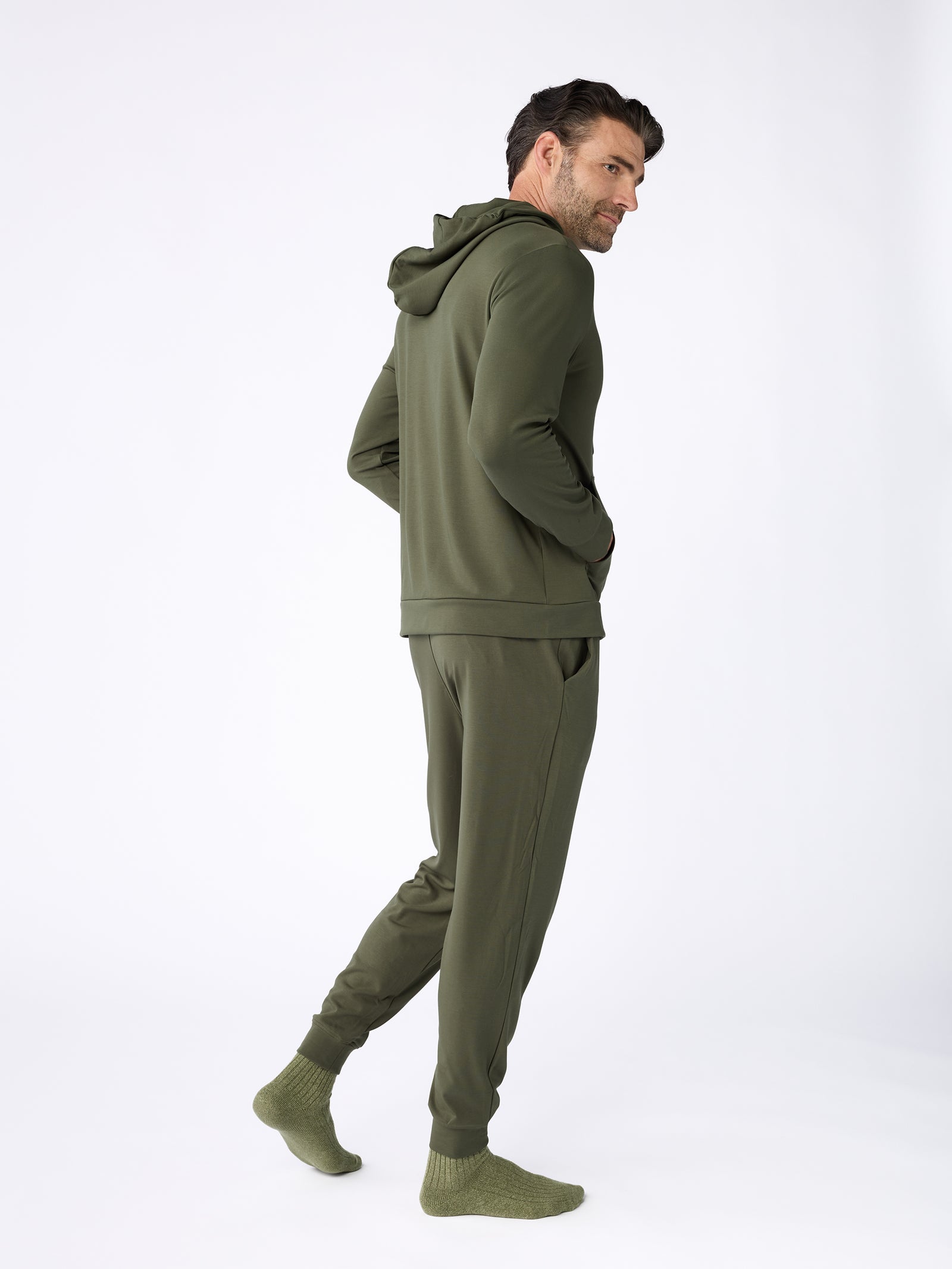 A man wearing the Men's Ultra-Soft Bamboo Hoodie by Cozy Earth and matching joggers stands in a relaxed pose with his hands in his pockets. The background is plain white, and he is also wearing green socks. 