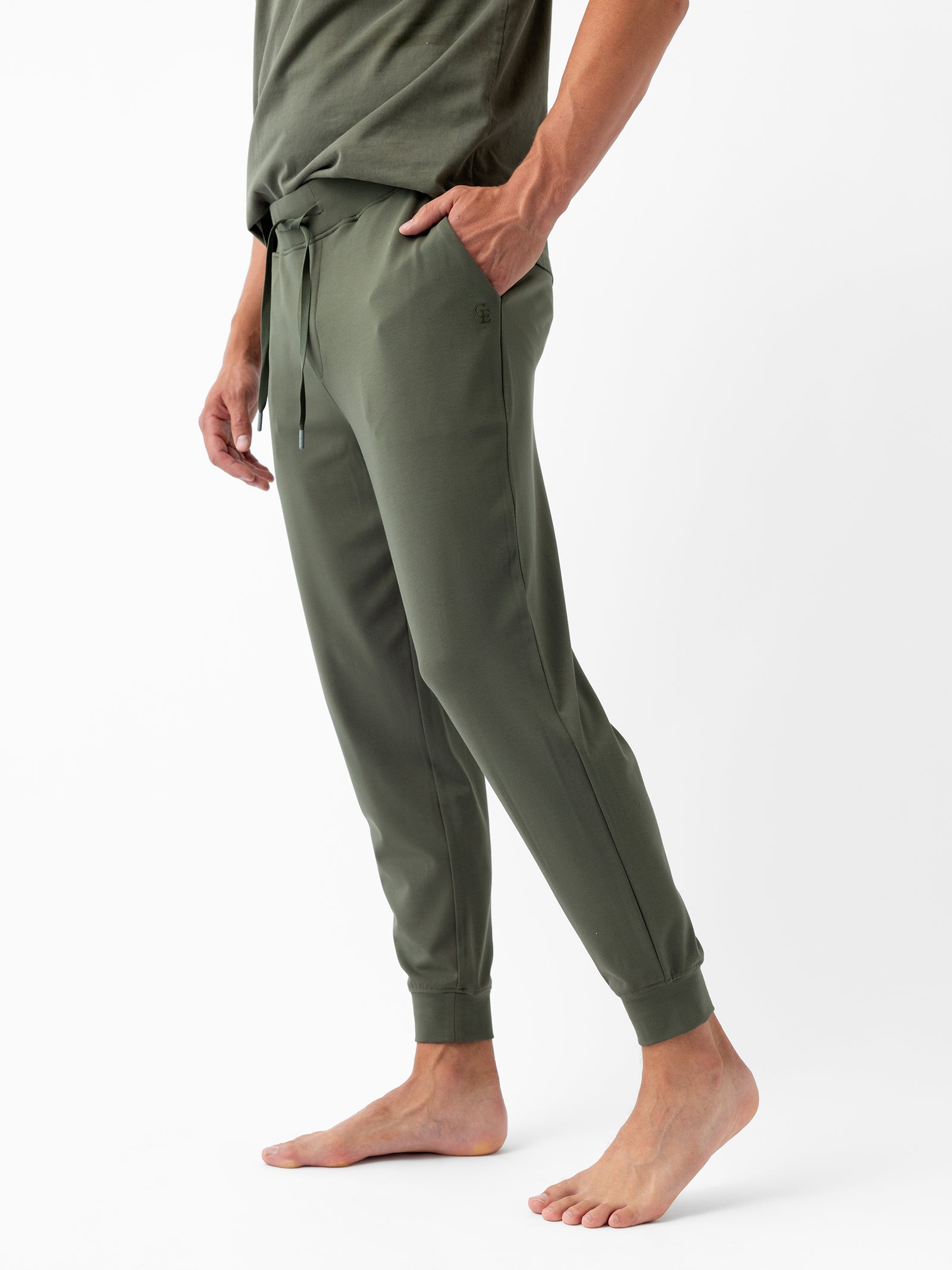 A person stands barefoot, adorned in Cozy Earth's Men's Ultra-Soft Bamboo Jogger Pant in a chic olive green hue, featuring a drawstring waist and practical pockets. These jogger pants elegantly taper at the ankles and are paired with a matching olive green shirt against a plain white backdrop. 