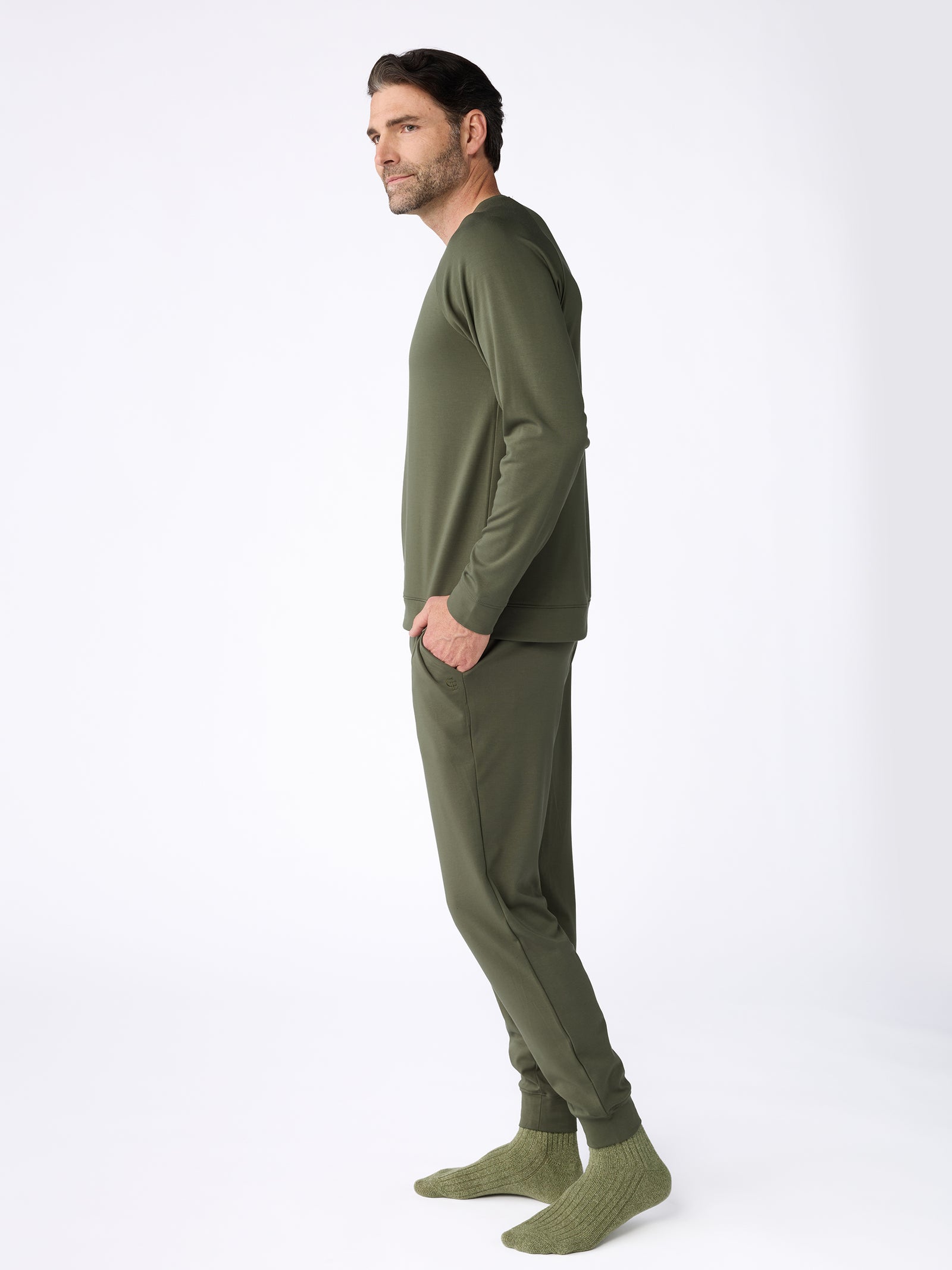 A man with dark hair and a beard stands in profile against a white background, wearing Cozy Earth's Men's Ultra-Soft Bamboo Pullover Crew in olive green, paired with matching joggers and knit socks, his hands tucked into the pockets of his pants. 