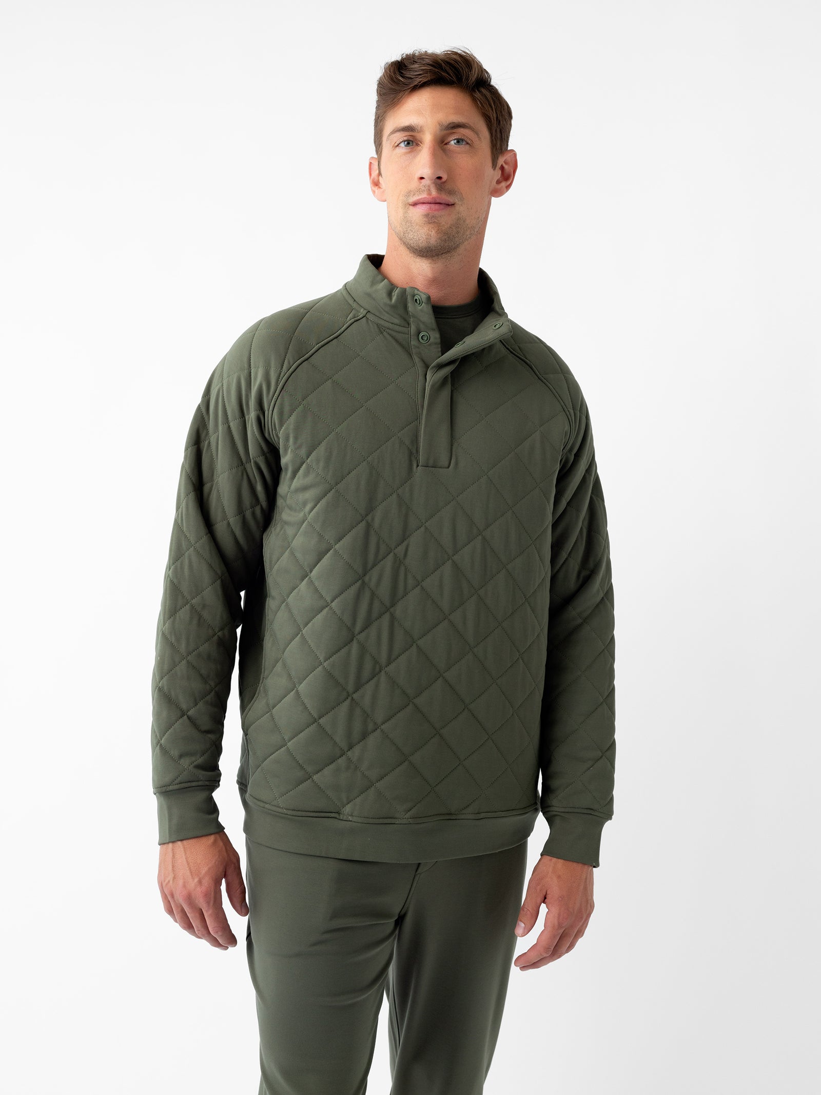 A person stands against a plain white background, donning the Men's Ultra-Soft Bamboo Quilted Snap Pullover from Cozy Earth. The green pullover features a high collar and snap-button closure. The individual, with short light brown hair, looks directly at the camera with a relaxed and confident posture. 