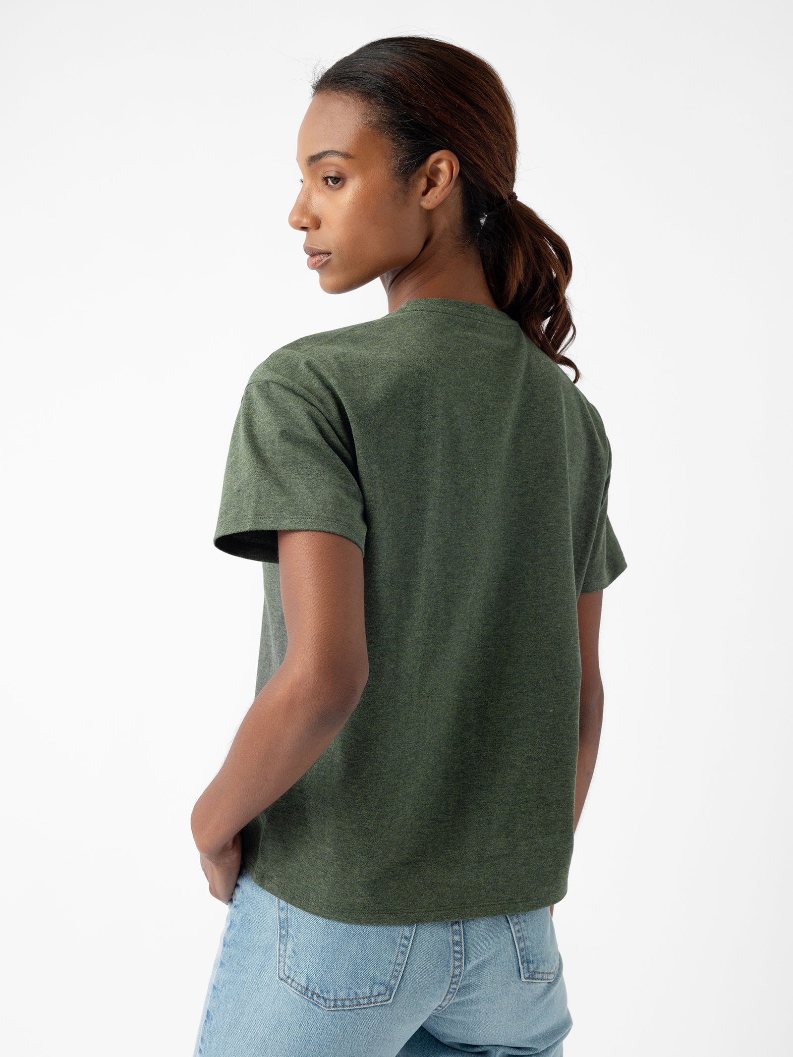 A person with dark hair in a ponytail stands facing away from the camera, looking over their left shoulder. They are wearing the Cozy Earth Women's All Day Tee in green and light blue jeans. The background is plain white. 