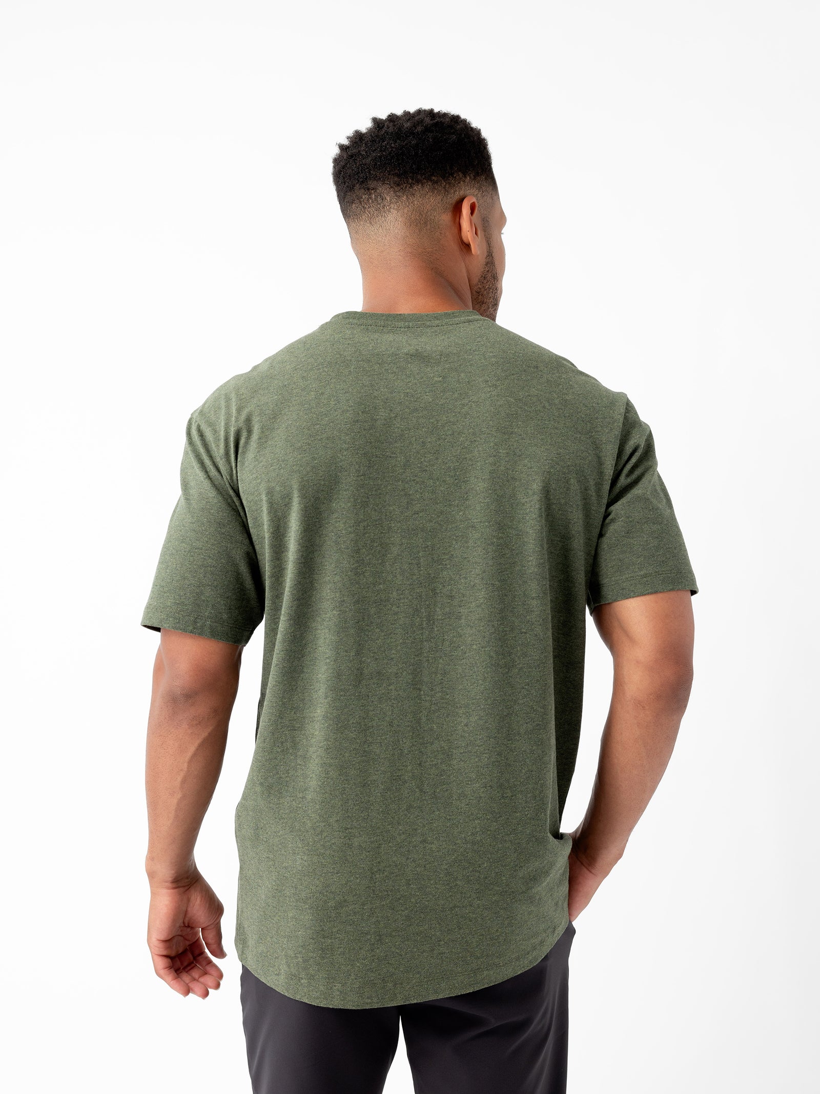 A person with short black hair and a trimmed beard is facing away from the camera, wearing a short-sleeved, green Men's All Day Tee by Cozy Earth and dark pants. They are standing against a white background. 