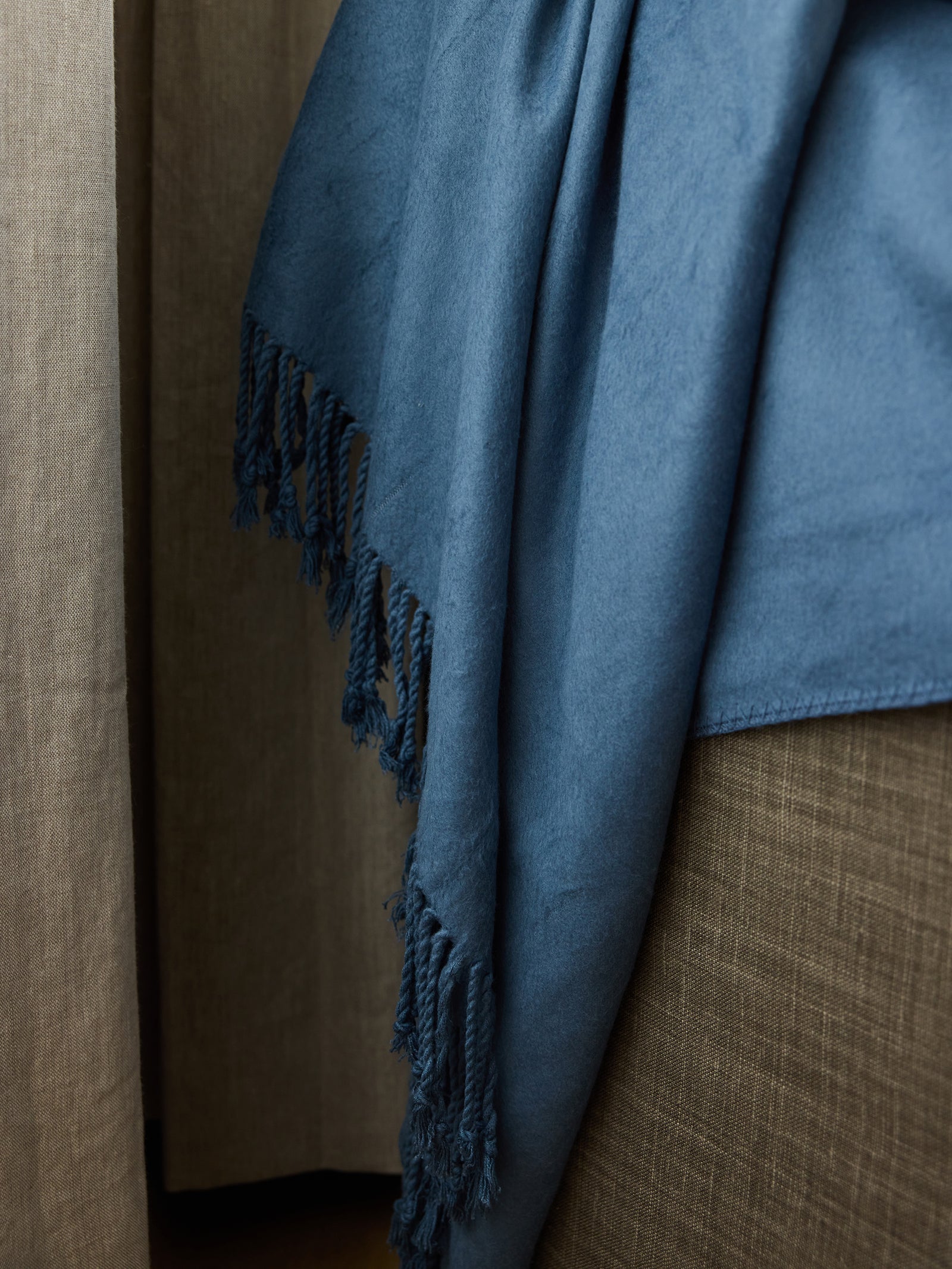 A close-up image of Cozy Earth's Bamboo Tassel Throw in blue, featuring intricate fringes, hanging over a brown textured surface. The throw contrasts against the brown background, showcasing its soft texture and detailed design. 