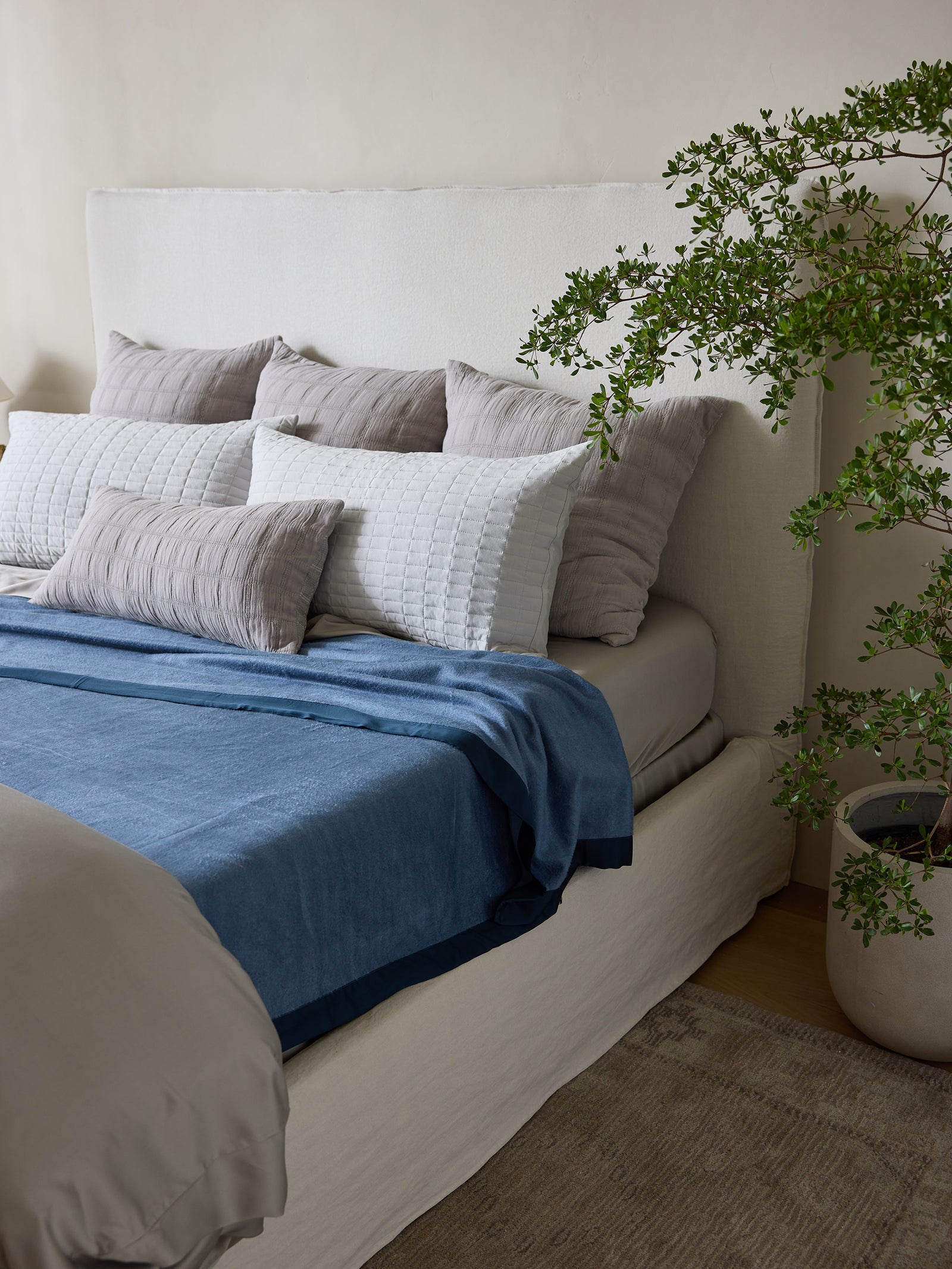 A cozy bedroom features a neatly made bed with a white headboard and multiple pillows in white and gray hues. A Bamboo Blanket by Cozy Earth, in a soothing blue shade, is layered on the bed. A green potted plant is placed nearby, adding a natural element to the neutral-toned room. 