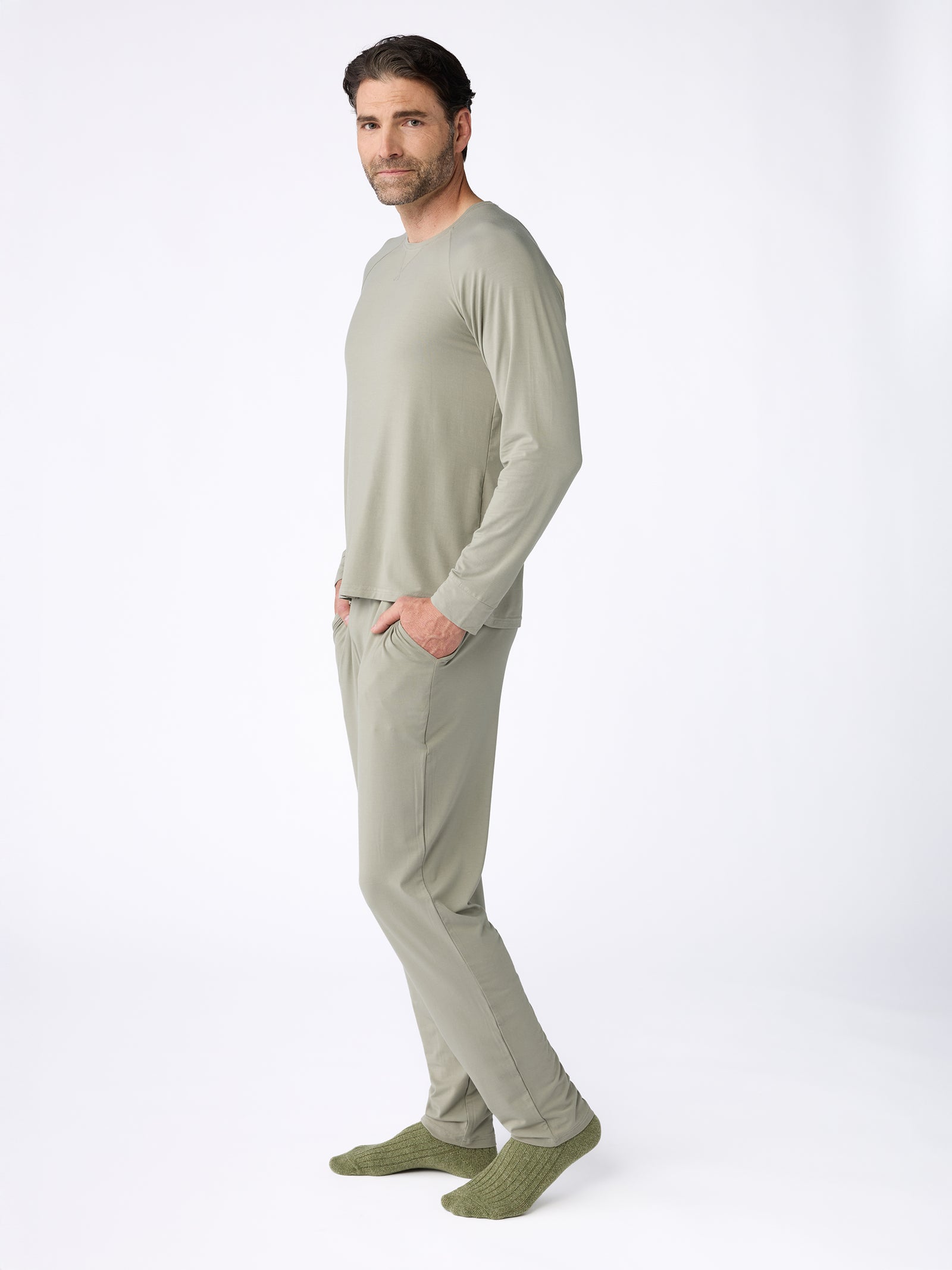 A person wearing the Men's Stretch-Knit Bamboo Long Sleeve by Cozy Earth in a light grayish-green hue, along with matching pants and green socks, stands against a plain white background. With their hands in their pockets, they gaze slightly to their left. 