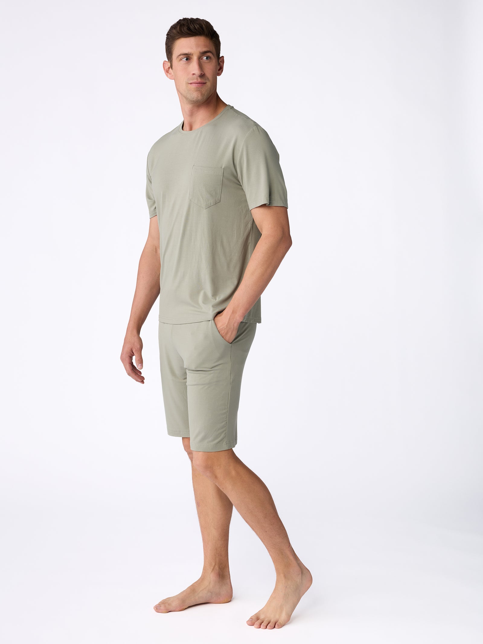 A man stands barefoot wearing a light green t-shirt and a pair of Cozy Earth's Men's Stretch-Knit Bamboo Pajama Shorts. The t-shirt features a pocket, and the outfit exudes a casual and comfortable vibe against the plain white background. 