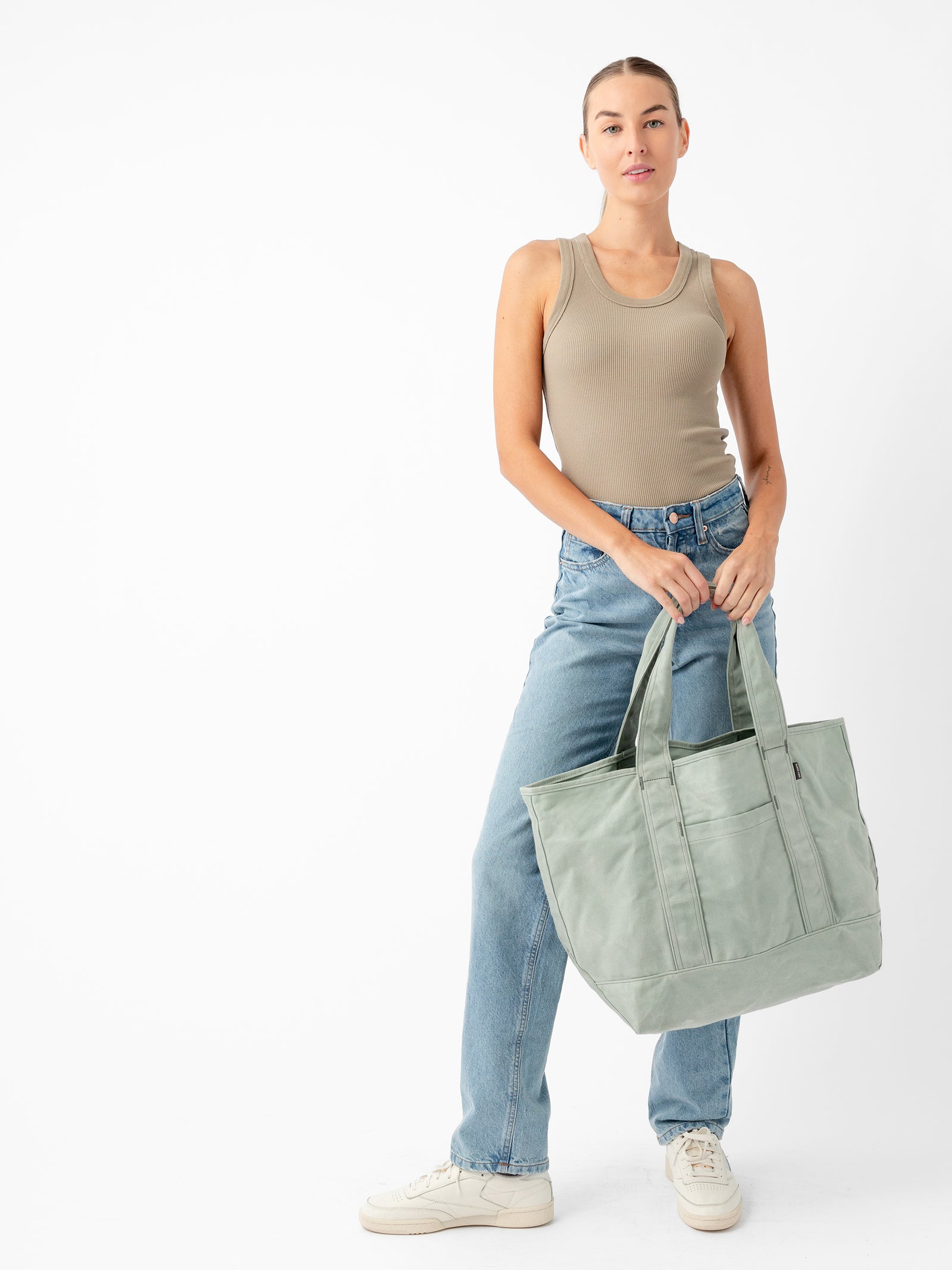 A person with their hair in a bun stands against a white background. They are wearing a beige sleeveless top, blue jeans, and white sneakers. They are holding the Cozy Earth Waxed Canvas Tote in light green with both hands in front of them. 