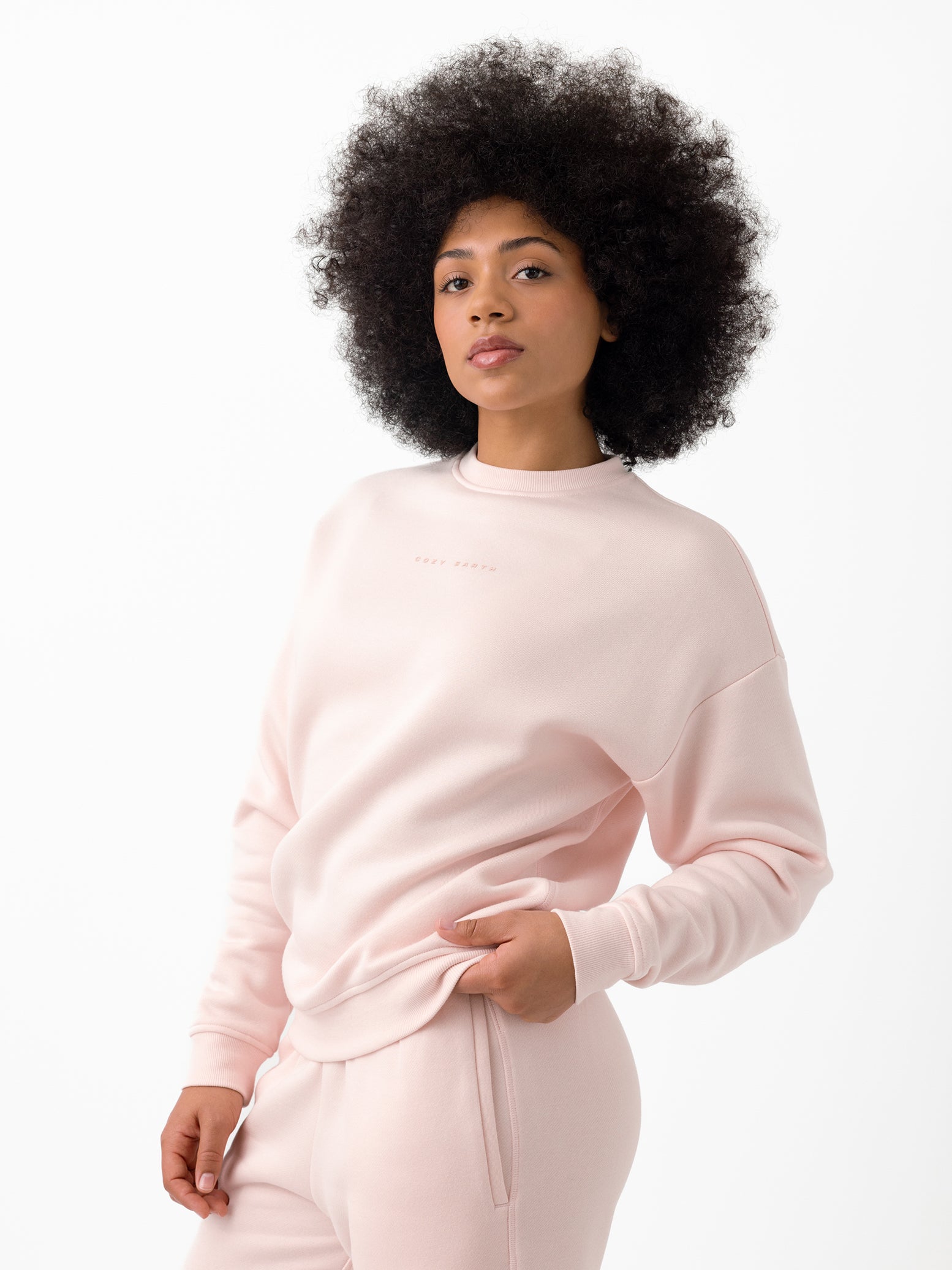 A person with curly hair stands against a white background, wearing Cozy Earth's Women's CityScape Crewneck and matching pants. They have one hand in their pocket and a relaxed expression. 