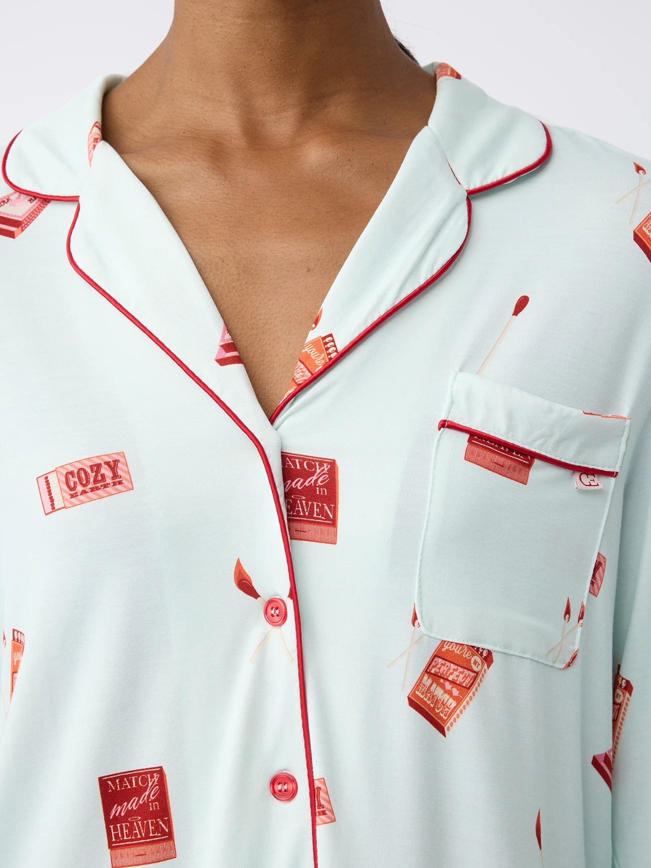 A woman wearing Cozy Earth's Women’s Stretch-Knit Long Sleeve Bamboo Pajama Set in light blue with red piping, buttons, and a chest pocket has a matchbox print reading "Cozy" and "Match Made in Heaven. 