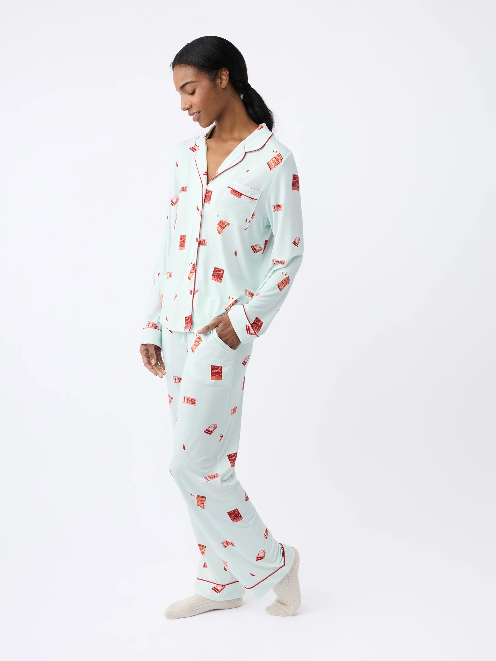 A woman stands sideways against a white background, wearing Cozy Earth Women's Long Sleeve Bamboo Pajama Top in Stretch-Knit, light blue with a red pattern. She has slippers on and appears relaxed as she looks down. 