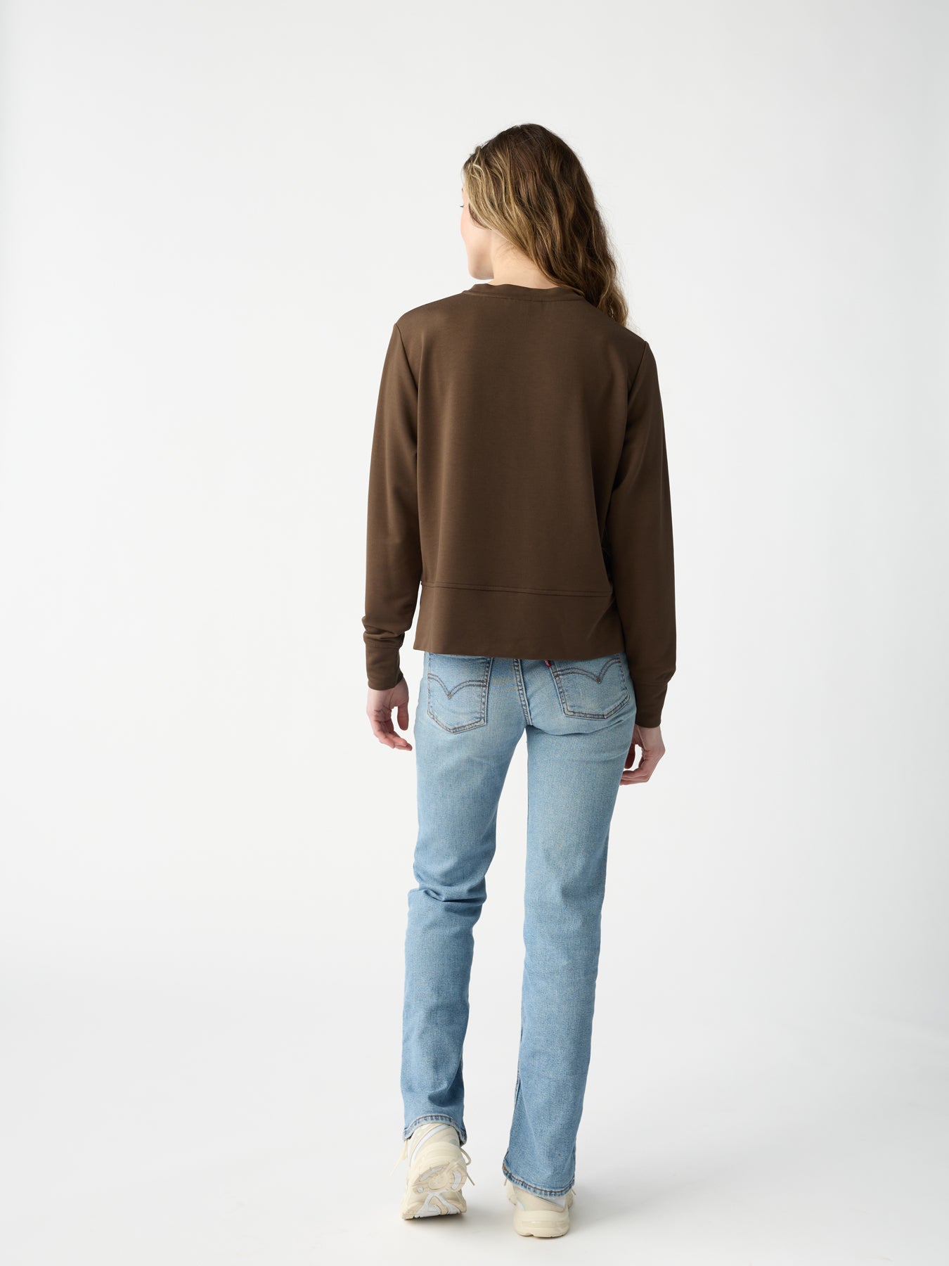 A person with long hair wearing Cozy Earth's Women's StudioLite Crewneck and light blue jeans stands against a plain white background, facing away from the camera. 