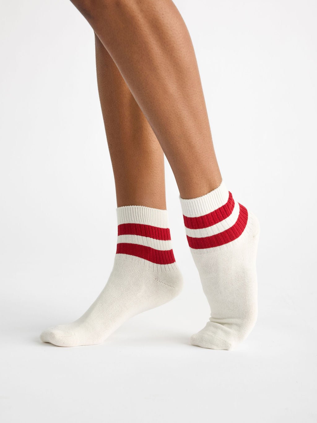 A person wearing Cozy Earth's Women's Sadie Sock, featuring a white design with two red stripes, balances on one foot against a plain white background. 