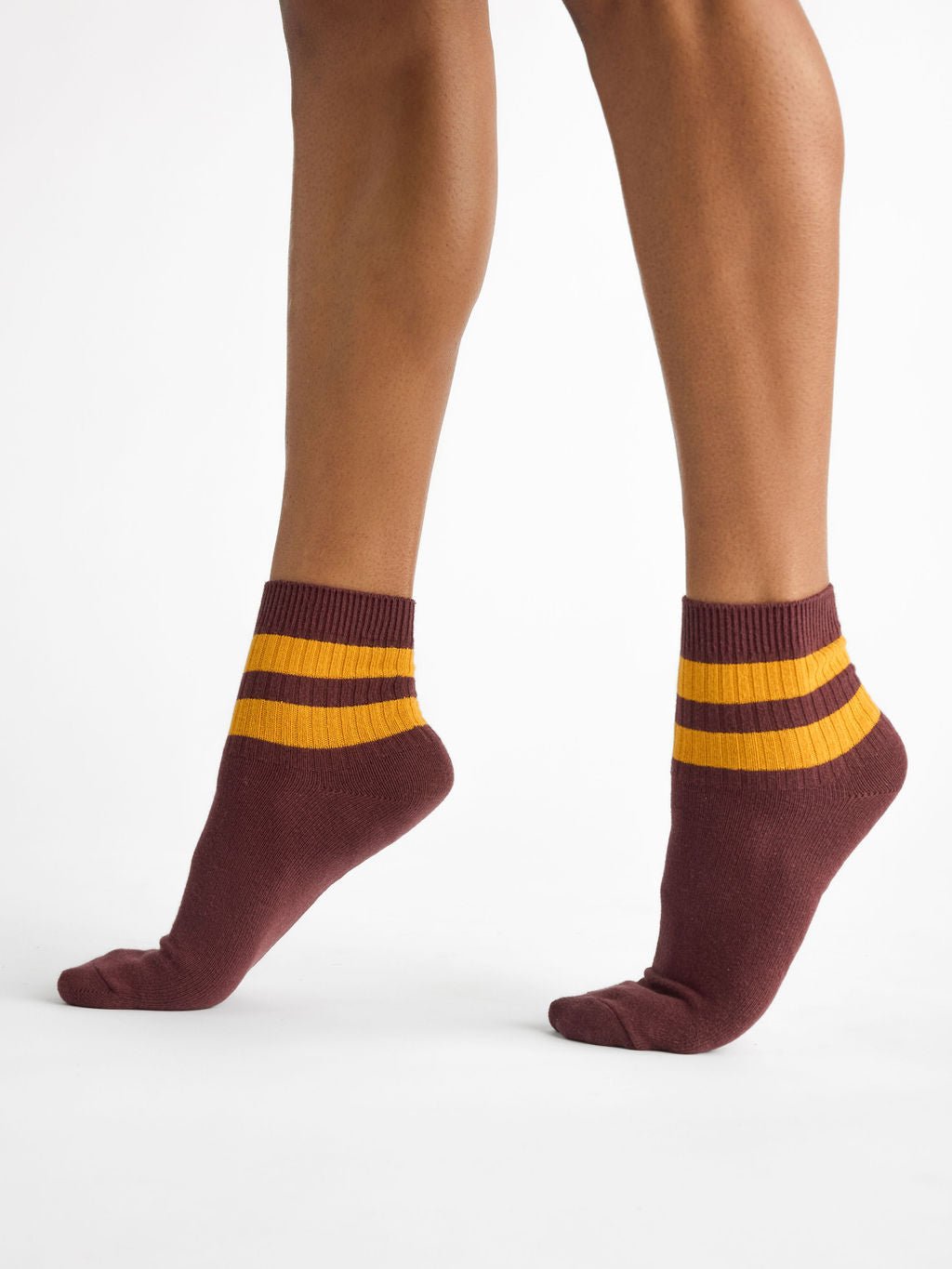 A person is wearing the Women's Sadie Sock by Cozy Earth, featuring a maroon color with two horizontal mustard-yellow stripes. They are standing to display the side profile of their feet and lower legs against a plain white background. 