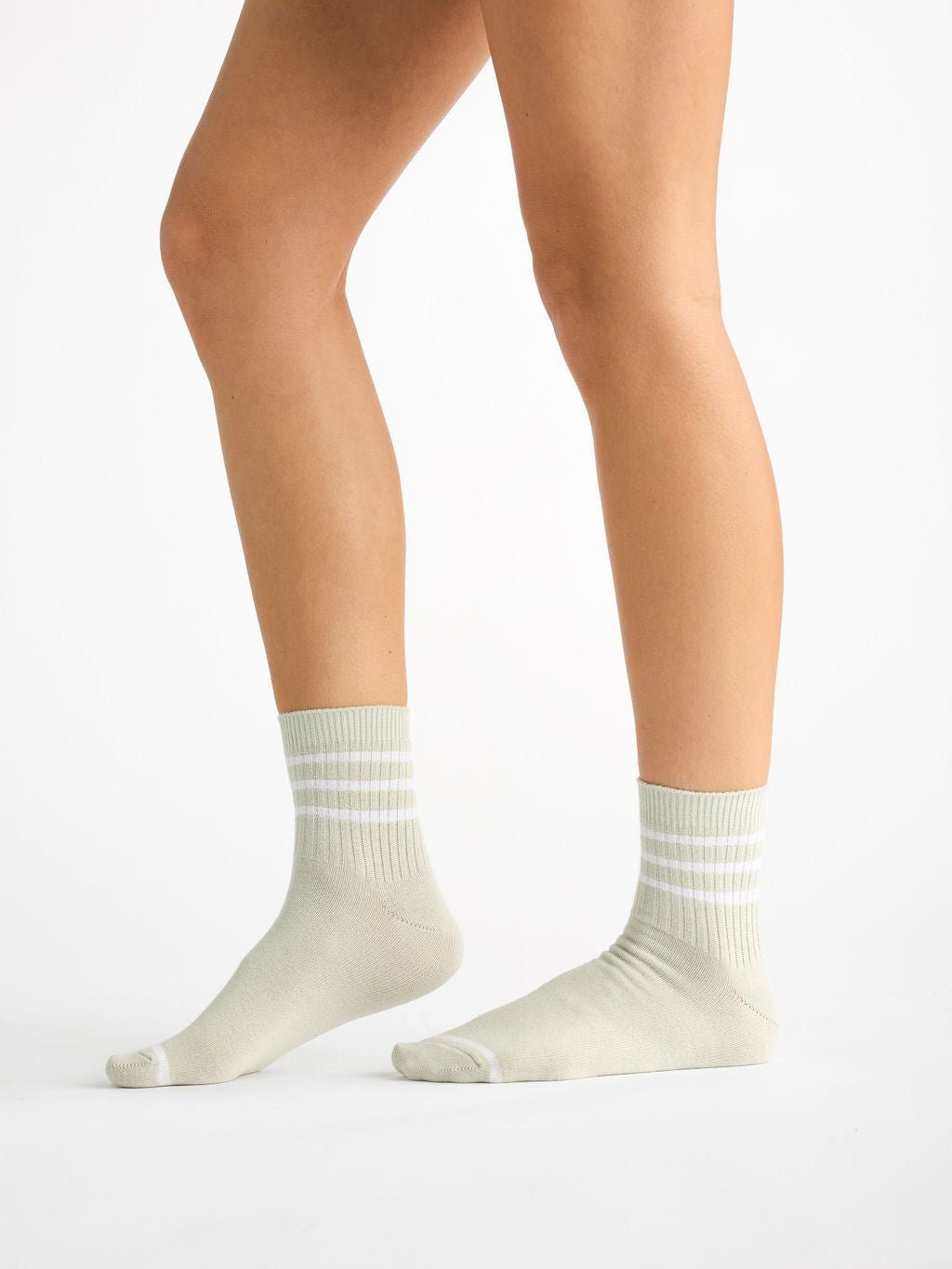 On a white background, a person wearing the mid-calf length, snug-fitting Women's Sadie Socks by Cozy Earth is shown from the thigh down. The socks are light-colored with white stripes. 