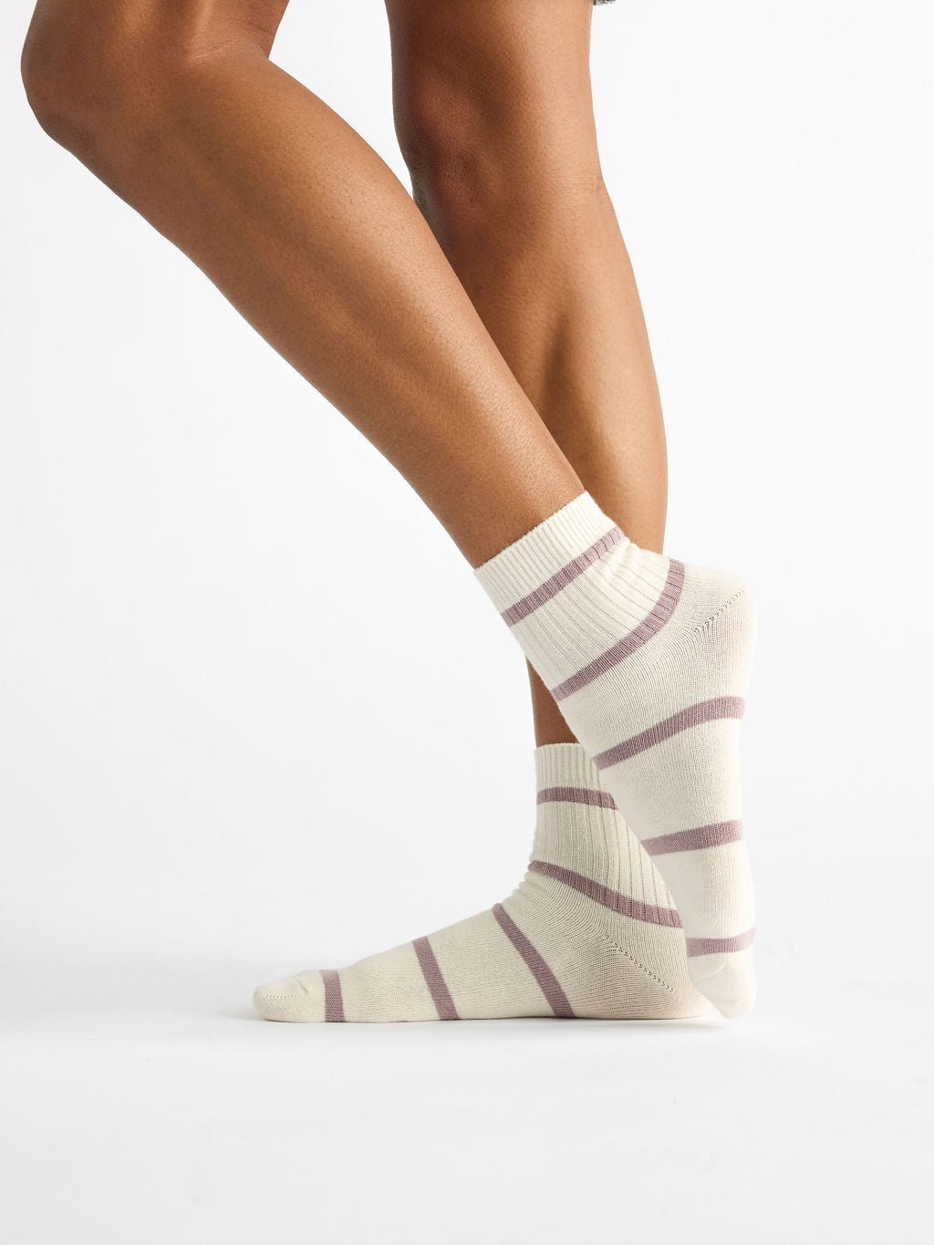 A person wearing Women's Sadie Sock from Cozy Earth, featuring a cream color with purple stripes, is standing on one foot with the heel of the other foot raised against a plain white background. 