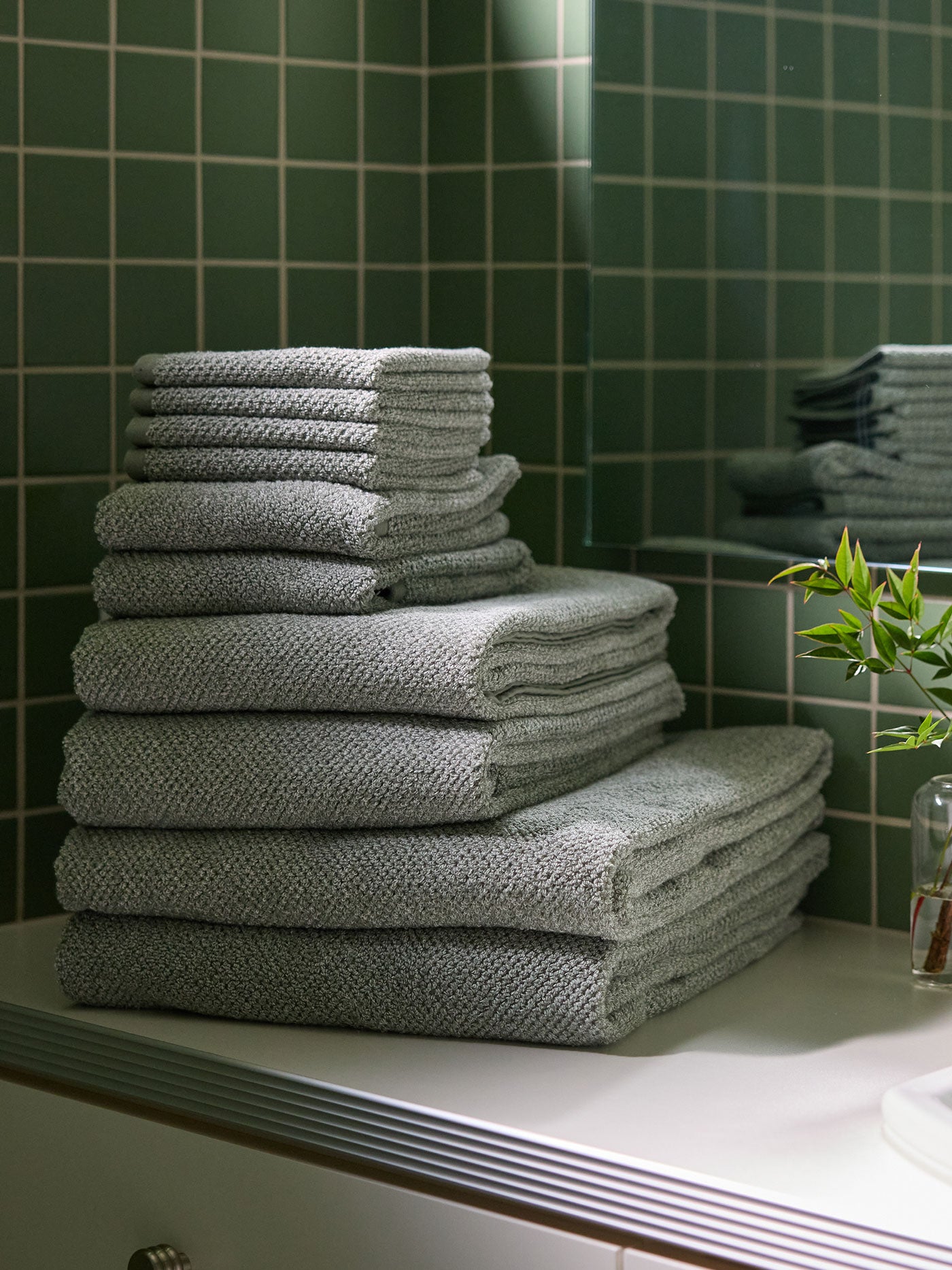 A stack of Nantucket towels in various sizes sit on the counter of a bathroom vanity. The wall behind is tiled in green tile. |Color:Heathered Sage