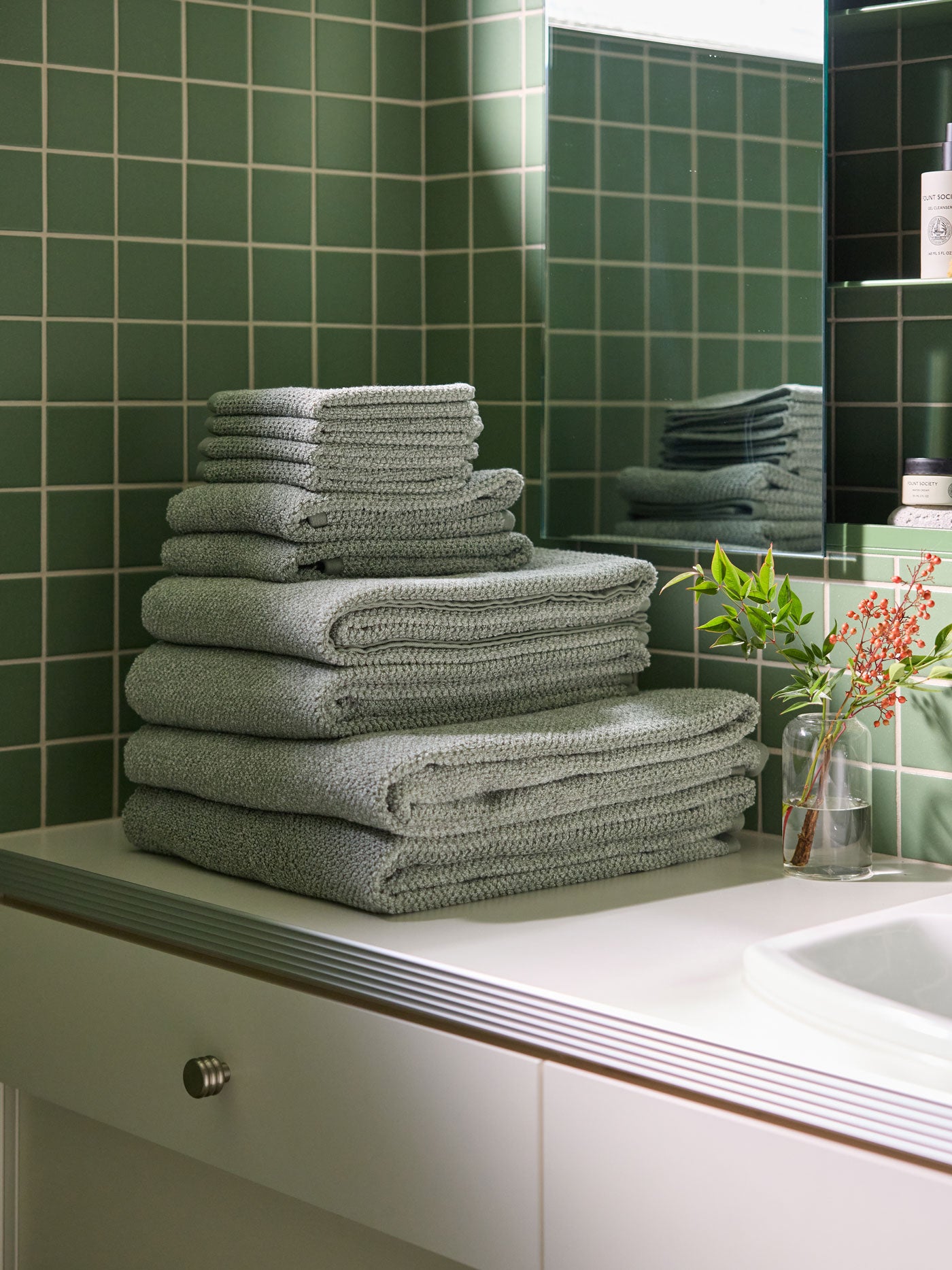 A stack of Nantucket towels and washcloths in Heathered Sage sit on a bathroom vanity countertop. There is a green tiled wall and shelves of skincare products in the background. 