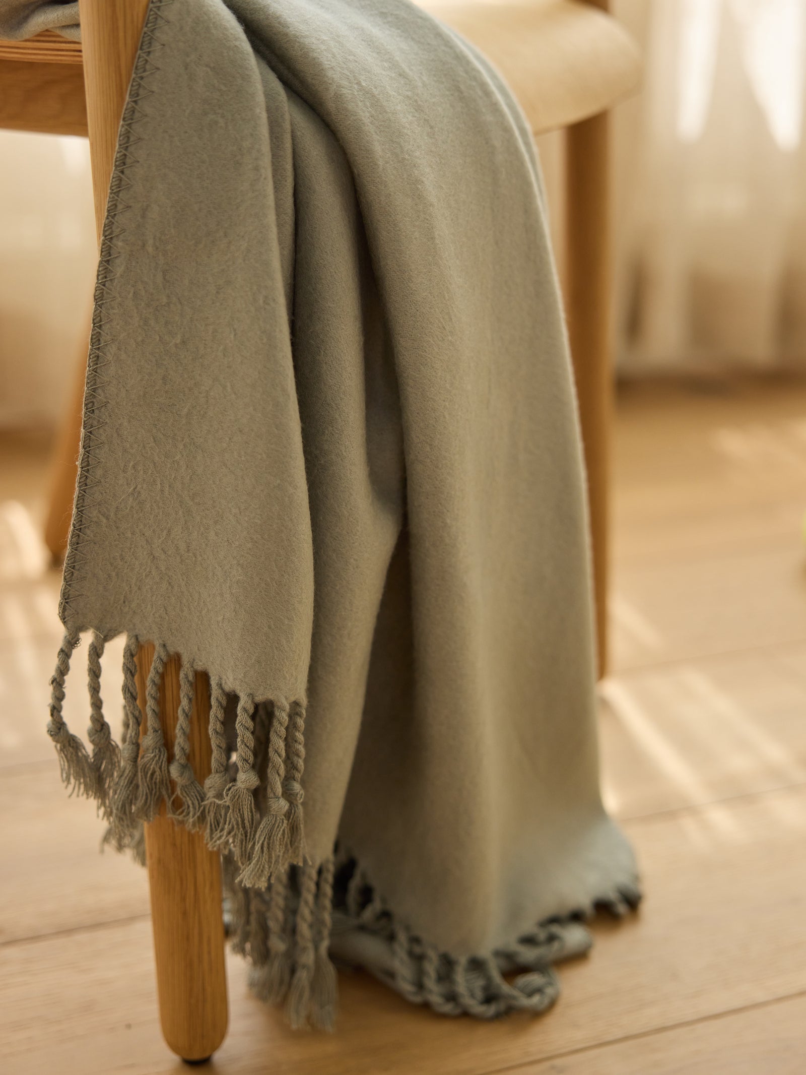 A Bamboo Tassel Throw by Cozy Earth, light gray with fringe edges, is draped over the back of a wooden chair. Sunlight casts soft shadows across the wooden floor and sheer curtains in the background, creating a warm and cozy ambiance. 