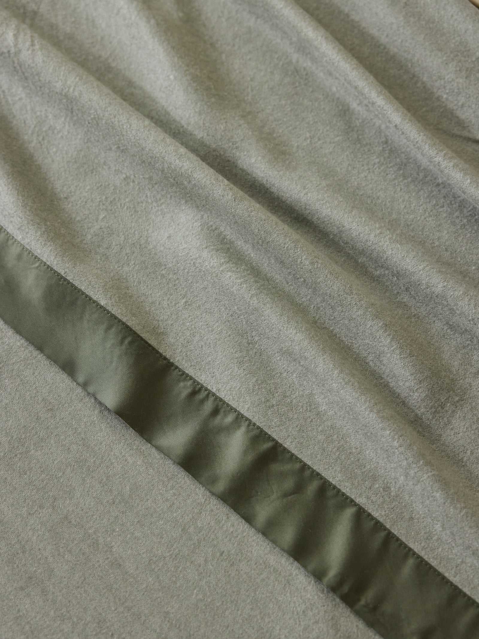 A close-up image of Cozy Earth's Bamboo Blanket reveals a textured fabric in a soft grayish-green color. A shiny, darker green satin stripe runs diagonally across the material, creating a striking contrast between the different textures. The fabric appears smooth and slightly wrinkled, emphasizing its elegance and comfort. 