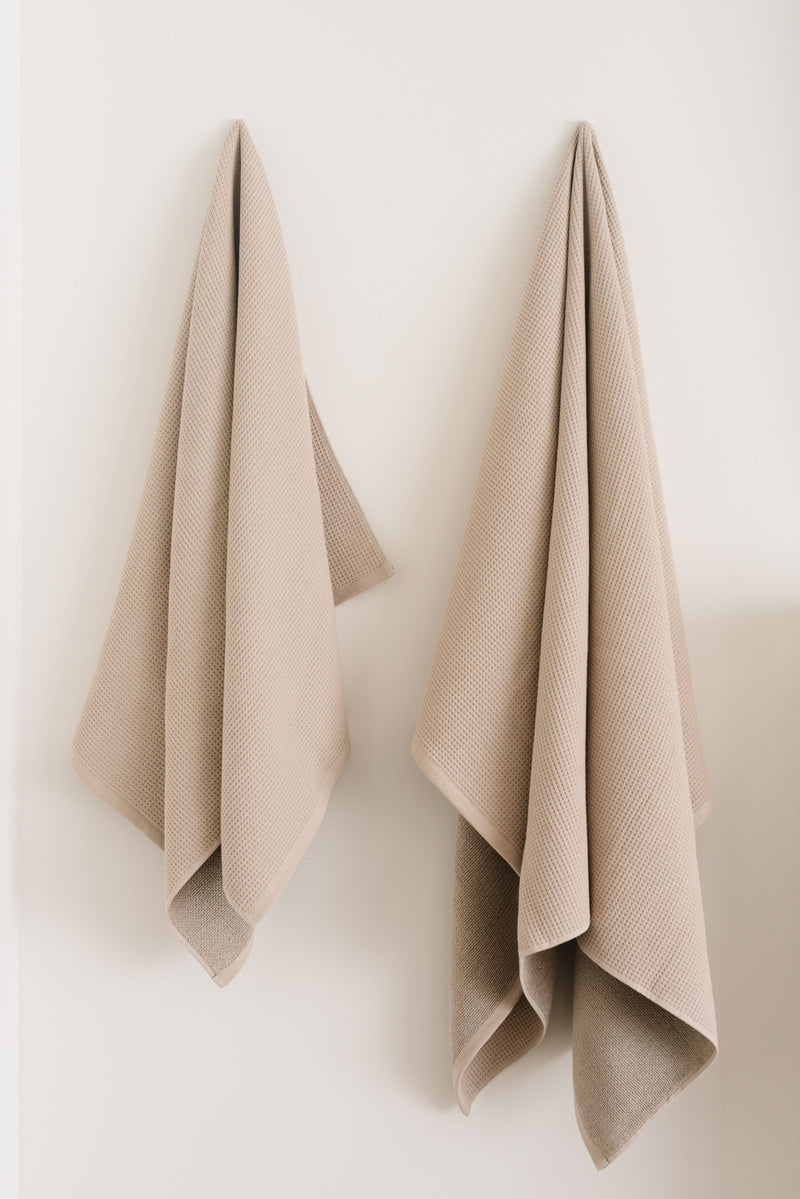 Waffle Bath Towel in the color Sand. Photo of Sand Waffle Bath Towel taken hanging from a towel hook in a white bathroom. The towel hook is not seen.