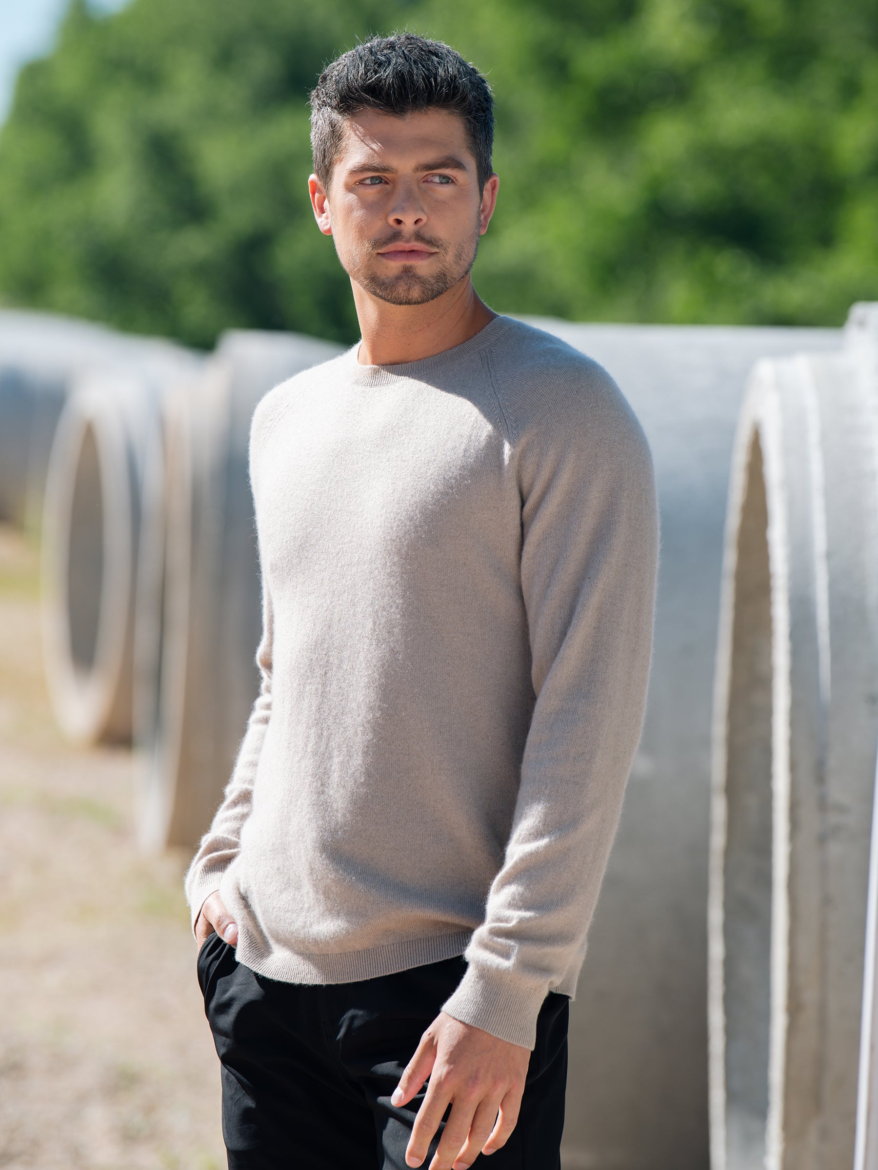 A man with short, dark hair and a trimmed beard is standing outdoors, looking away from the camera. He is wearing a light beige Men's Crewneck Sweater from Cozy Earth and black pants. Large cylindrical pipes are in the blurred background, along with greenery. |Color:Sandstone