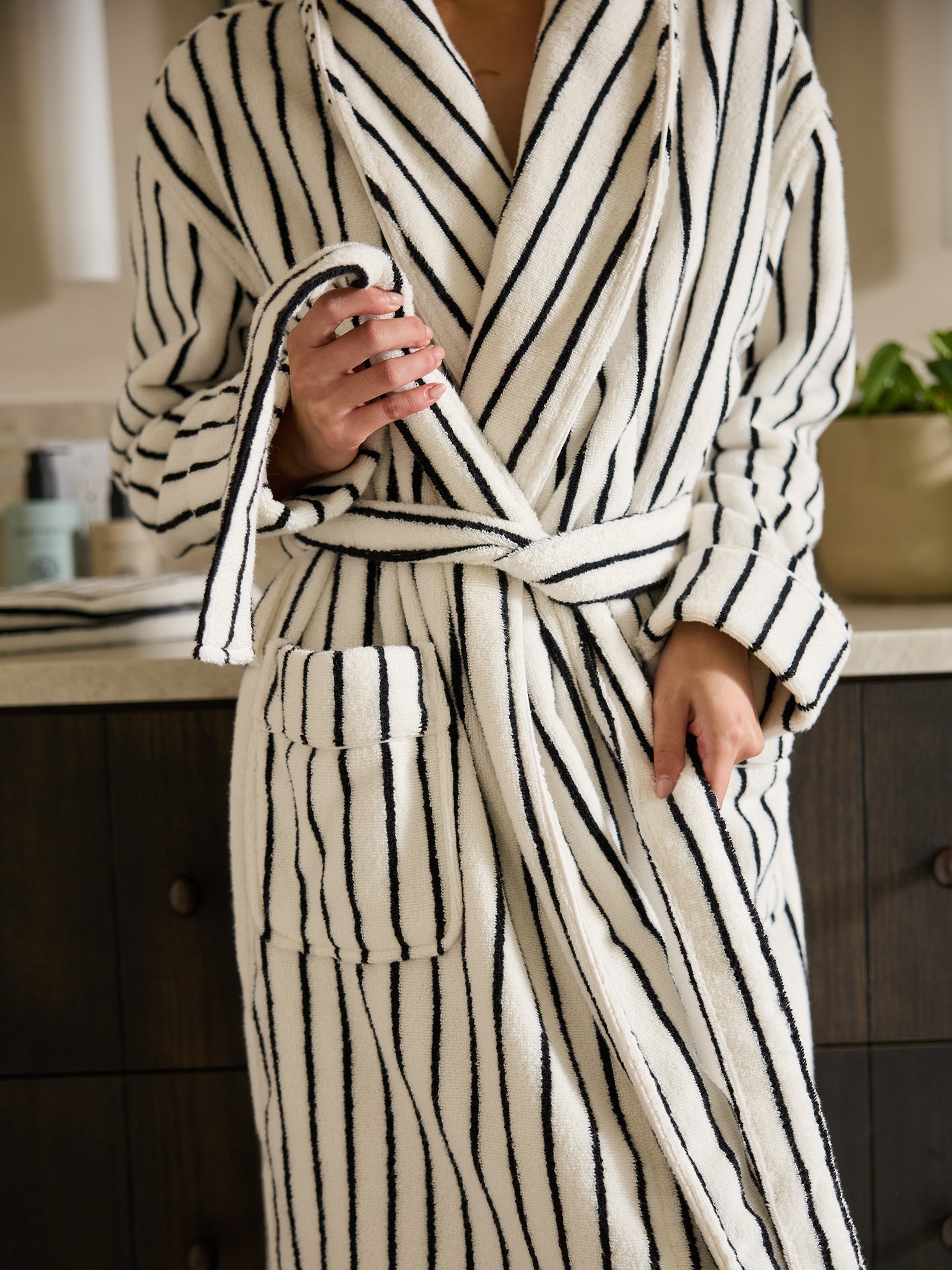 A person dressed in the Luxe Bath Robe from Cozy Earth, featuring black vertical stripes on a white background, stands beside a countertop adorned with a selection of toiletries and a potted plant. The person's hands are adjusting the robe's belt. 