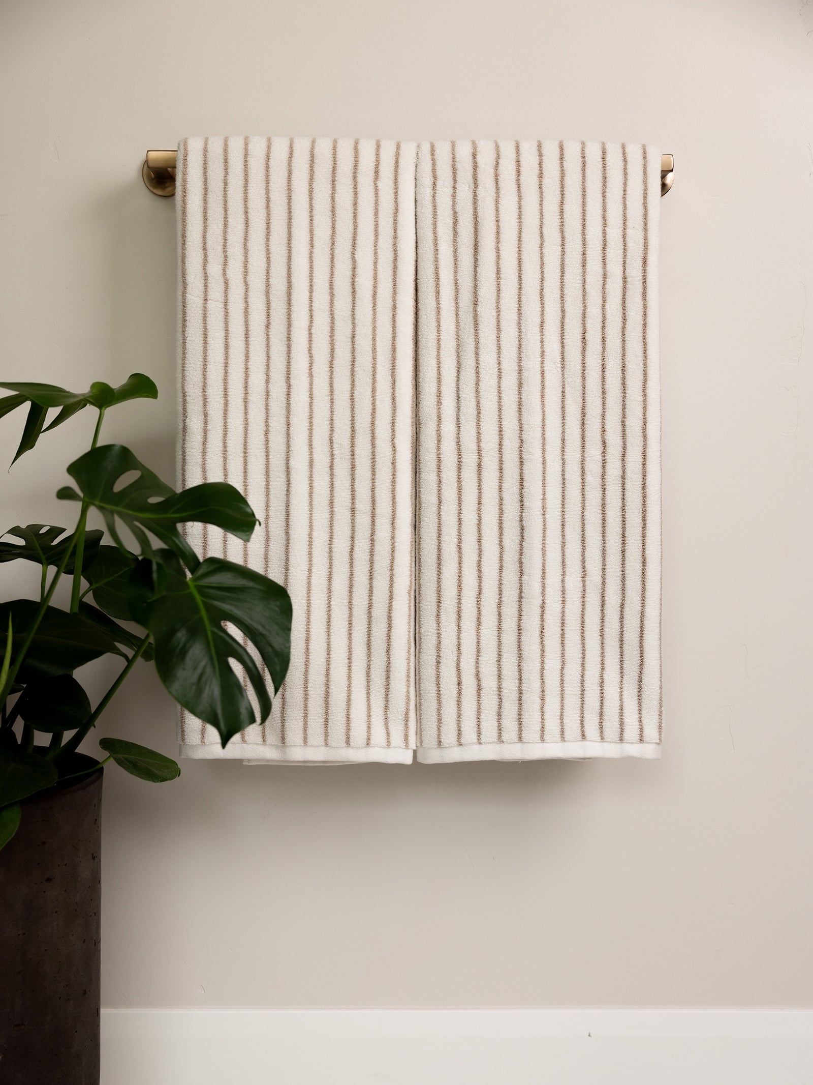 A Cozy Earth Luxe Bath Towel featuring thin, vertical beige stripes hangs neatly on a brass towel bar against a light, neutral-colored wall. To the left of the towel, a green potted plant with large leaves adds a natural touch. The overall scene is minimalist and calming. 