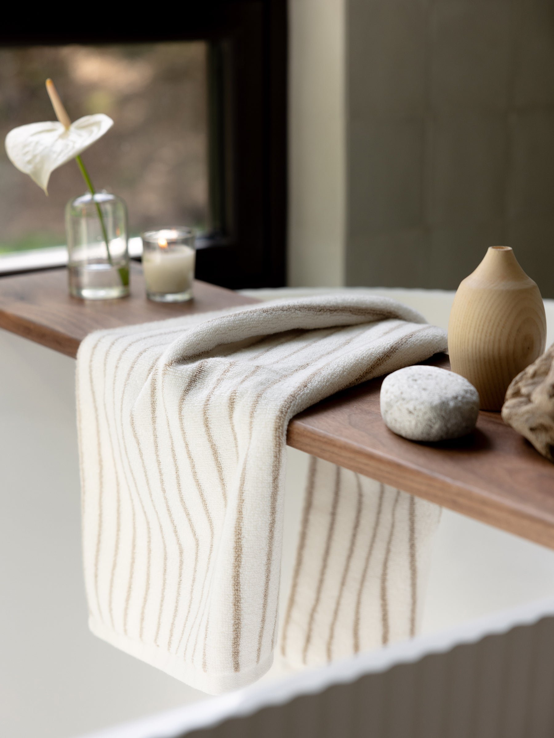 A Luxe Hand Towel by Cozy Earth, featuring a white and beige stripe pattern, hangs over the wooden ledge of a bathtub. Beside the towel on the ledge are a potted flower in a glass vase, a lit candle, a smooth stone, and a wooden decor piece. A window in the background offers a glimpse of greenery outside. 