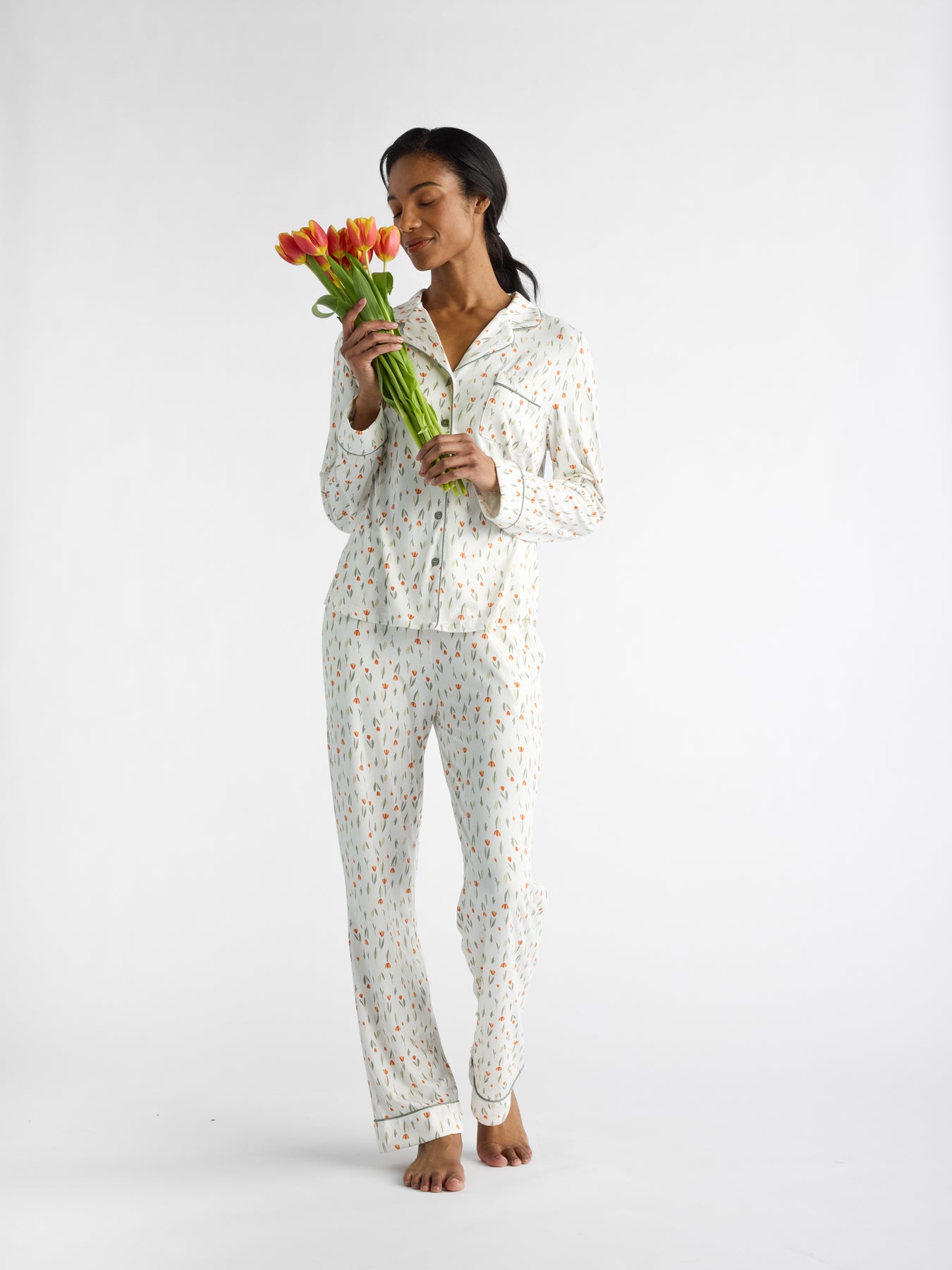 In a peaceful pose, a person in Cozy Earth's Women's Bamboo Stretch-Knit Long Sleeve Pajama Set stands barefoot, holding orange tulips with closed eyes against a plain white background. 