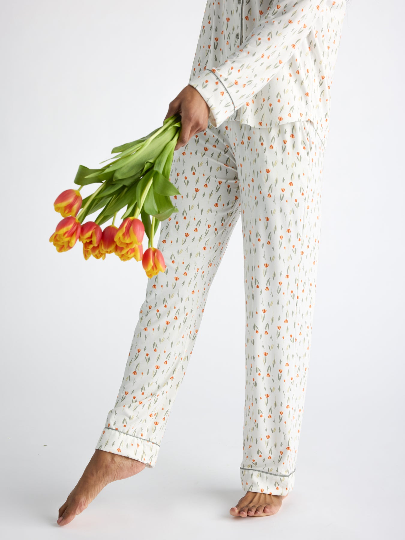 A person wearing Cozy Earth Women's Bamboo Stretch-Knit Pajama Pant holds orange and yellow tulips, standing barefoot against a white background. 