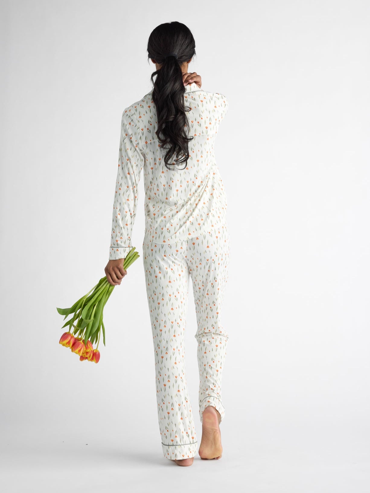 Wearing a Women's Bamboo Stretch-Knit Long Sleeve Pajama Top by Cozy Earth, a person with long hair walks barefoot away from the camera, holding orange tulips against a plain white background. 