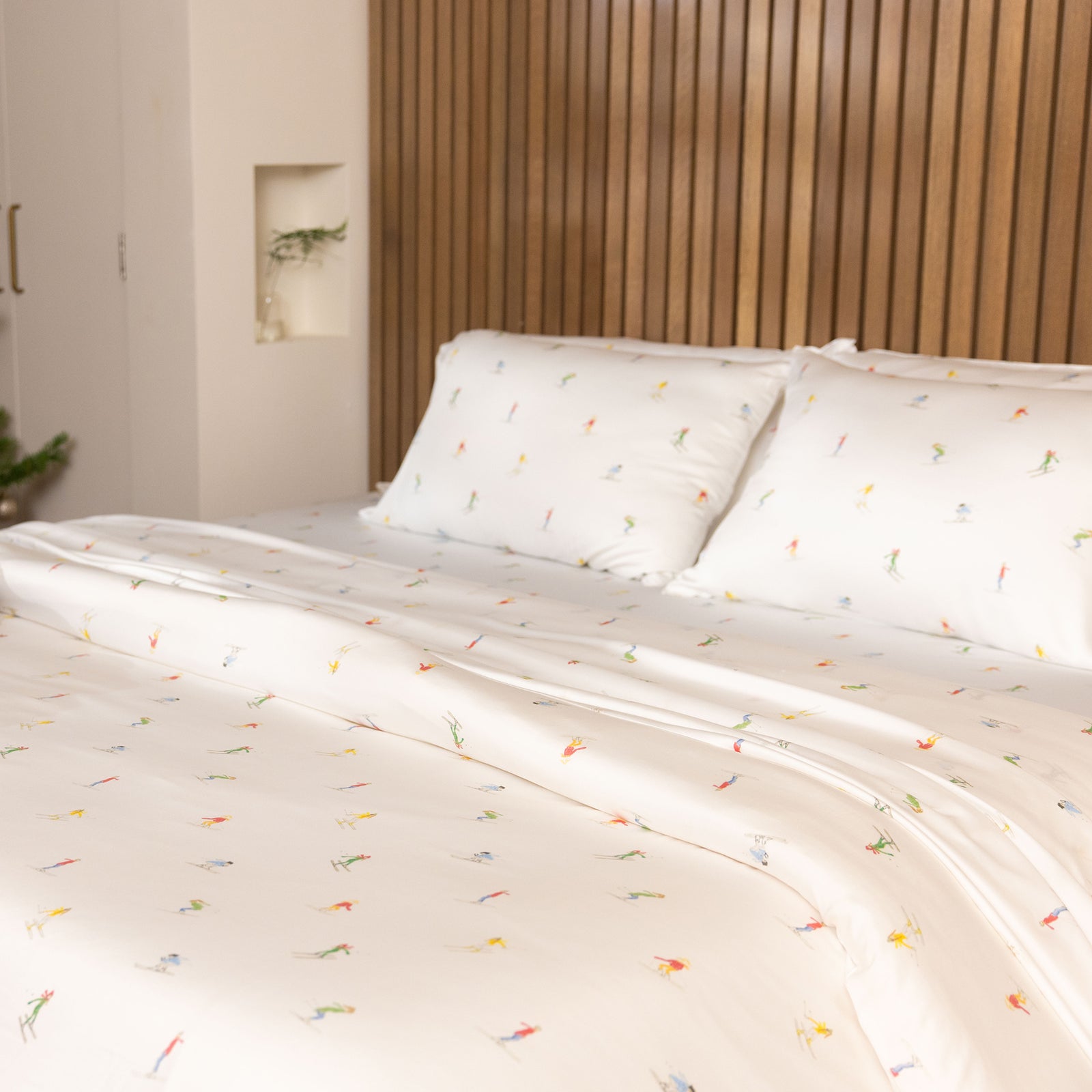 The bed is adorned with a Bamboo Duvet Cover from Cozy Earth, featuring white sheets and pillowcases with a subtle pattern of small, colorful figures. The headboard is crafted from vertical wooden slats, while soft lighting adds to the cozy ambiance. 