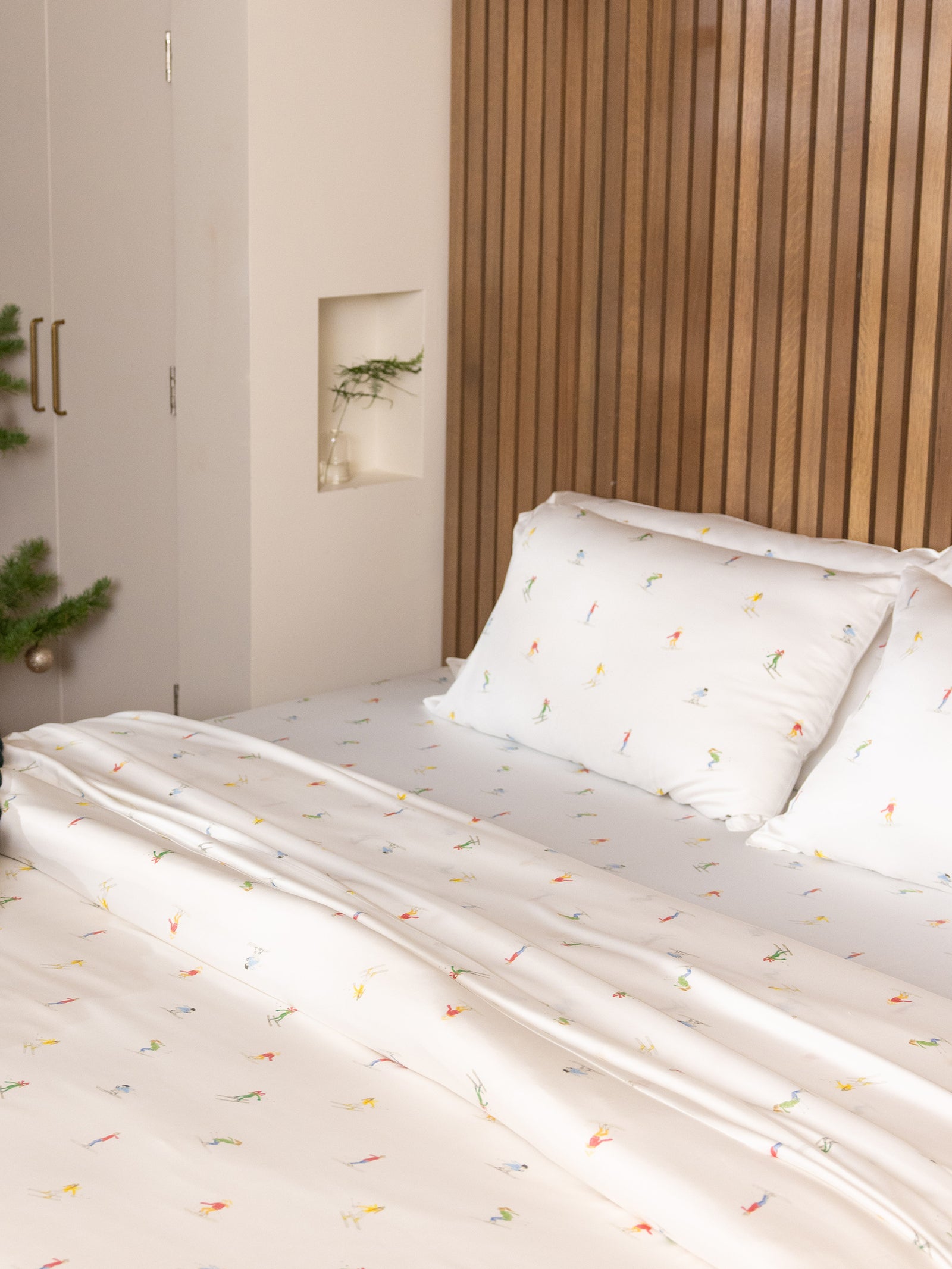 A neatly made bed with the Bamboo Sheet Set by Cozy Earth, featuring a small, colorful print. A wooden headboard and a small potted plant on a shelf are in the background, alongside a partial view of a closet with a stainless steel handle. 