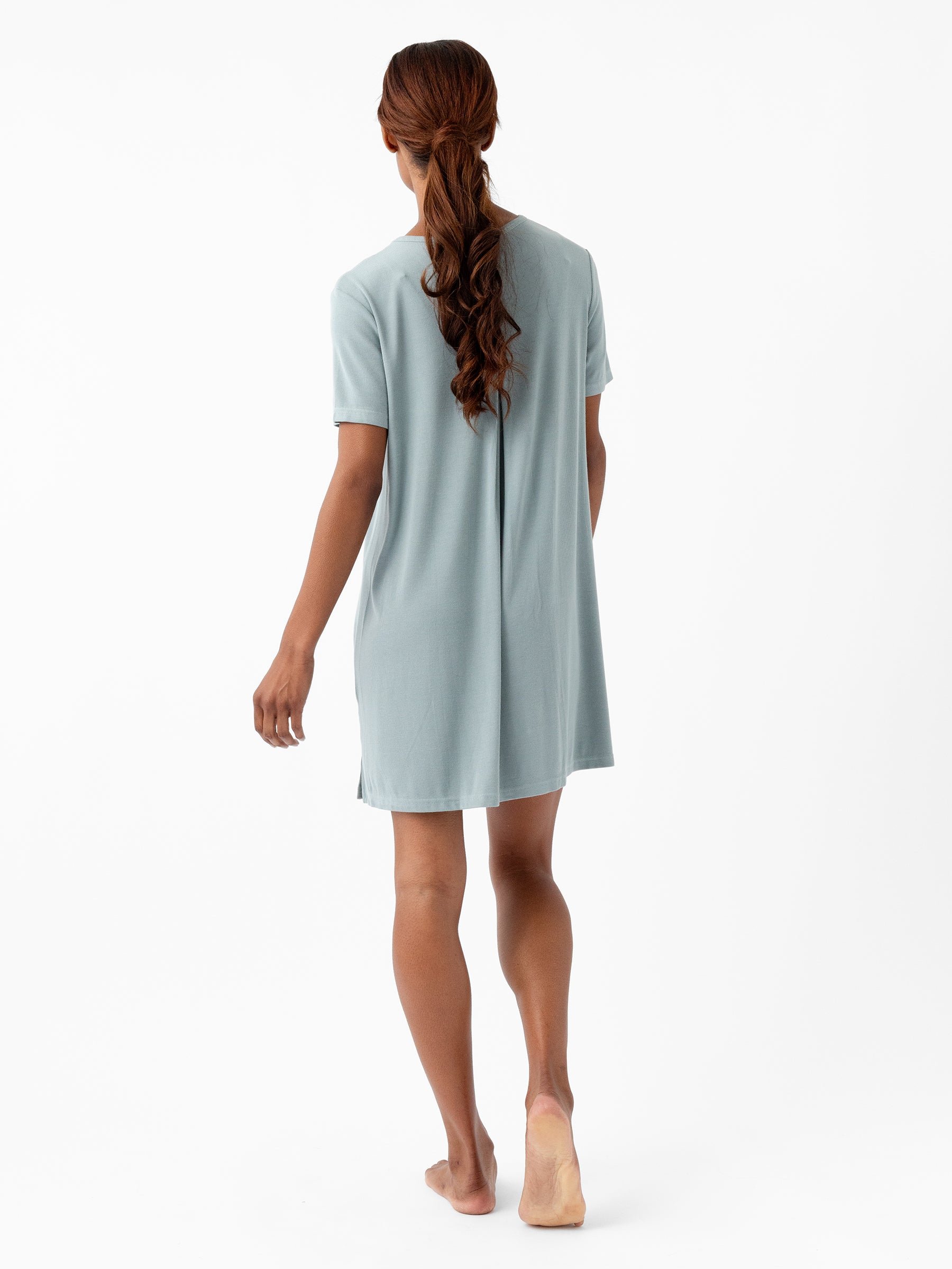 A person with long, wavy hair is shown from the back walking barefoot. They are wearing a Women's Bamboo Rib-Knit Boyfriend Sleep Dress by Cozy Earth in light blue, which has short sleeves and falls above the knees. The background is entirely white, emphasizing the subject and their attire. 