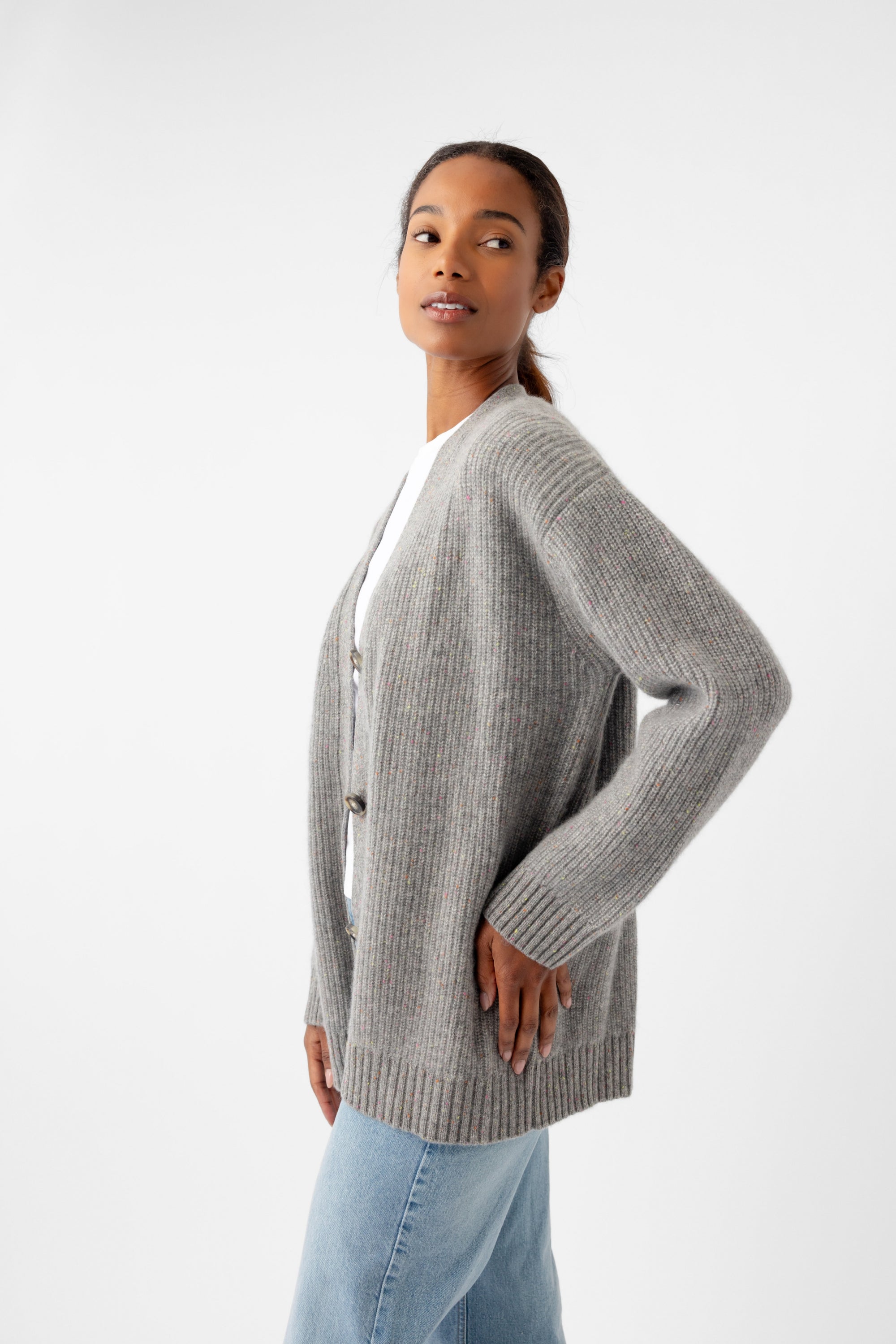 A person wearing an oversized Sunday Cashmere Cardigan from Cozy Earth in gray over a white shirt and light blue jeans stands against a plain white background, looking to the side with one hand on their hip. 